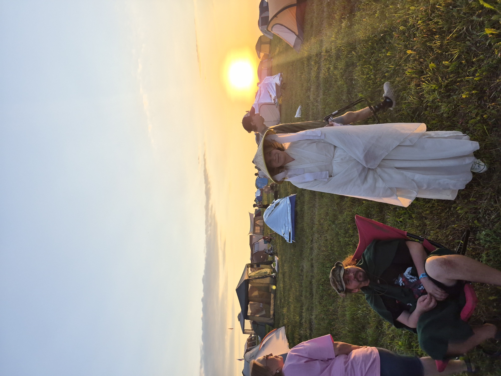 In Lagunovo, Russia, a campsite is illuminated by the setting sun, casting a warm glow over a field dotted with tents. In the foreground, a man sits in a red camping chair, wearing a camouflage cap and a green cloak. To his right, a person in a white robe and conical hat stands facing the camera, with a backdrop of tents and the setting sun. Another person, wearing a dark hat, stands behind the robed figure, looking towards the sun. A woman in a pink shirt is visible on the far left, her back to the camera. The environment is a grassy field with numerous tents of various shapes and sizes scattered across it.