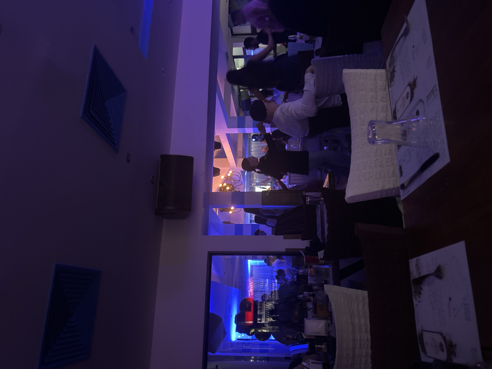 The image shows the interior of a restaurant in Plovdiv, Bulgaria. The room is lit with blue and purple lighting. Several people are seen standing and interacting within the space. Tables are set with napkins, menus, glasses, and silverware. A bar area with bottles and glassware is visible in the background. A speaker is mounted on the wall. Air vents are also visible on the wall.