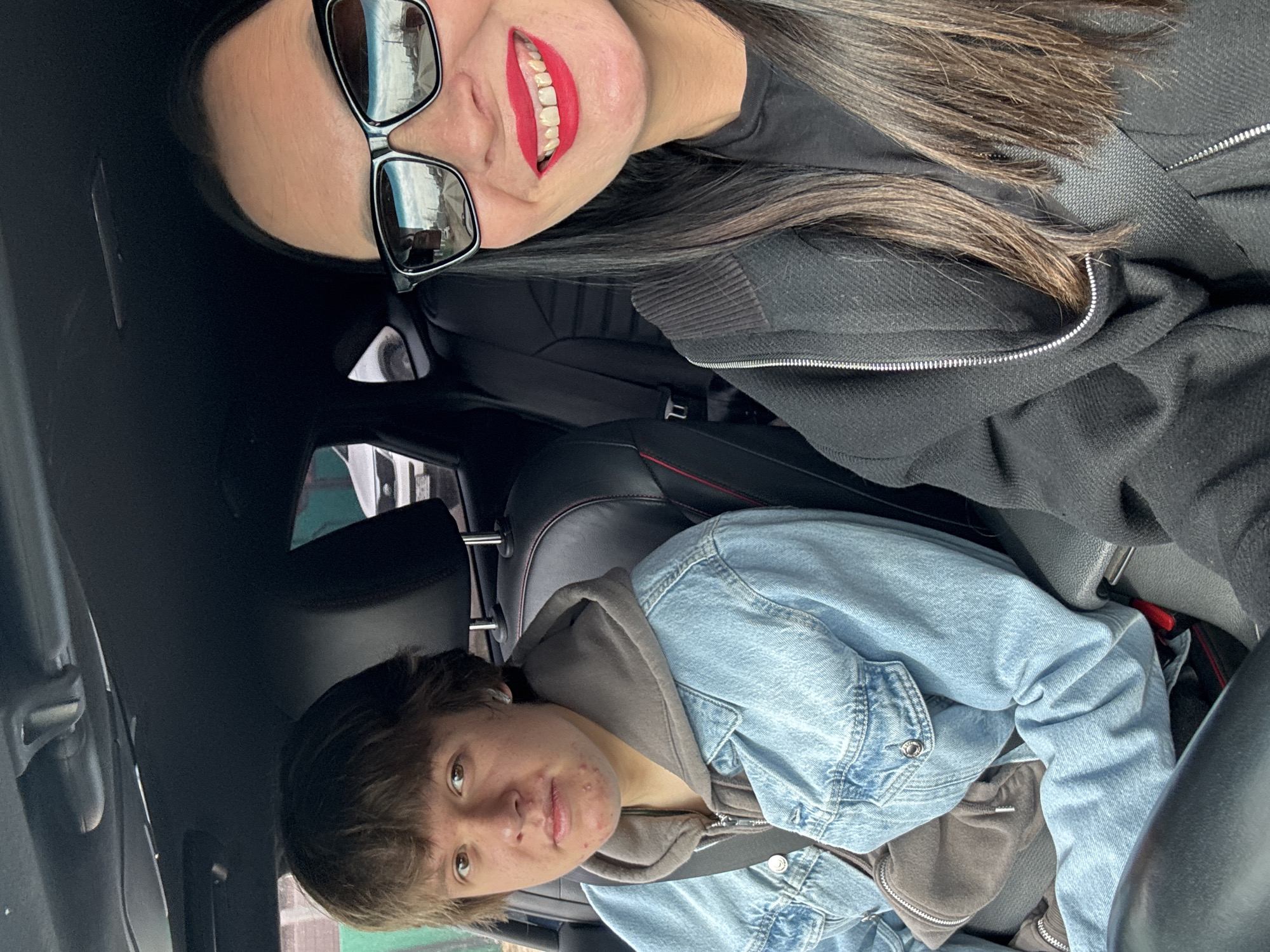 The image shows two people in the front seats of a car. A woman with long dark hair, wearing sunglasses and bright red lipstick, is in the passenger seat, smiling. She is wearing a black jacket and has a seatbelt on. In the driver's seat is a younger person with short brown hair, wearing a denim jacket over a hooded sweatshirt, and also wearing a seatbelt. The interior of the car is visible, including the seats, steering wheel, and dashboard. The location cannot be determined from the image and there is no indication that it is in the city of Chaikovsky, Russia.