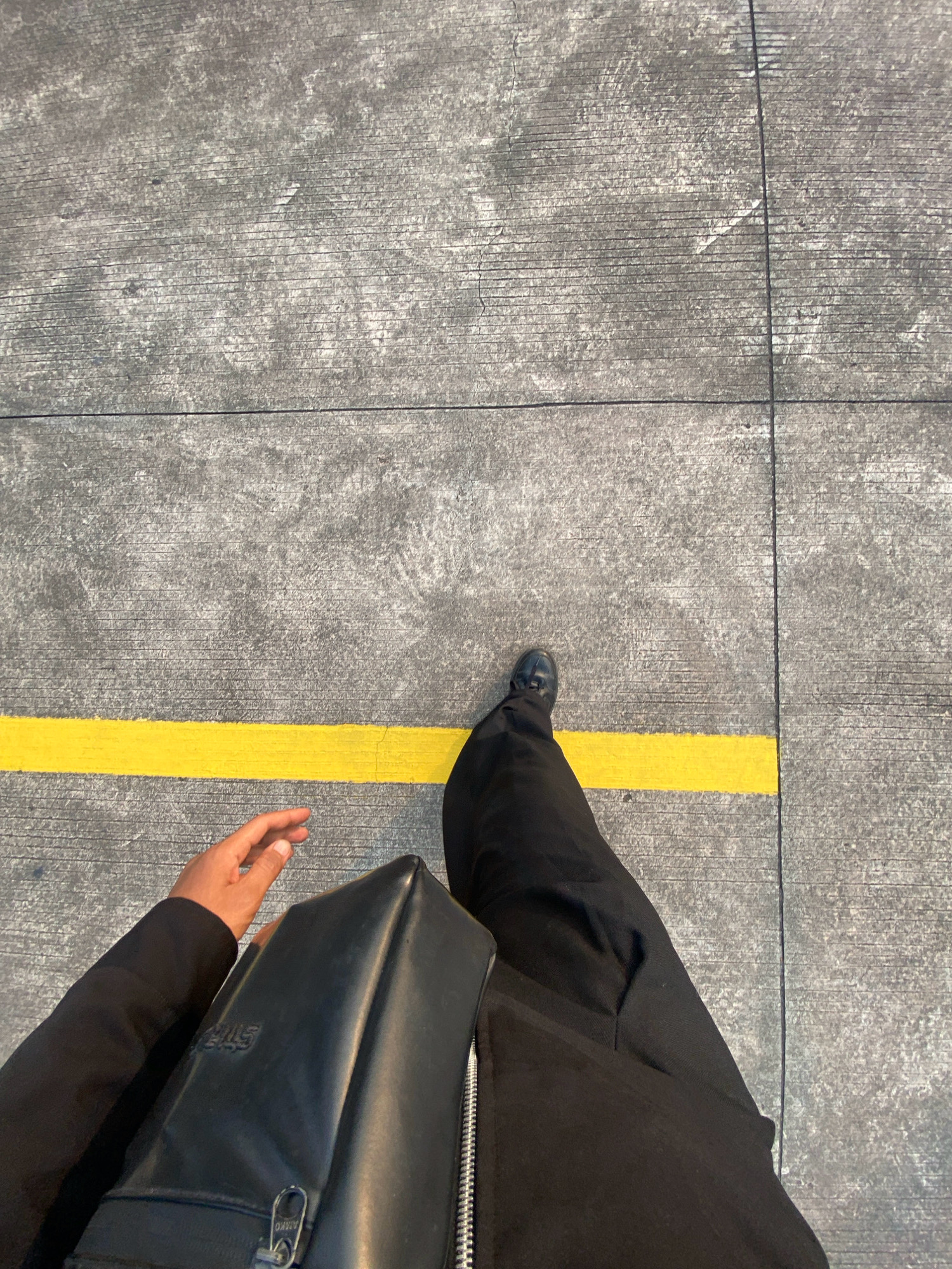 A first-person perspective shot shows a person's left leg, clad in black trousers and a black dress shoe, stepping over a bright yellow line painted on a grey concrete surface. The concrete appears to be an outdoor area with visible lines and textures, possibly a pathway or part of a larger structure. In the lower left of the frame, a hand, with fingers slightly spread, is visible near a black bag. The bag, made of a smooth, shiny material, has a zipper and embossed lettering. The scene is lit by sunlight, casting shadows and highlighting the textures of the concrete. The image is taken at a downward angle. The yellow line and concrete texture suggest an area within Hang Nadim International Airport (BTH) in Batam, Indonesia, potentially near the tarmac or a designated walkway.