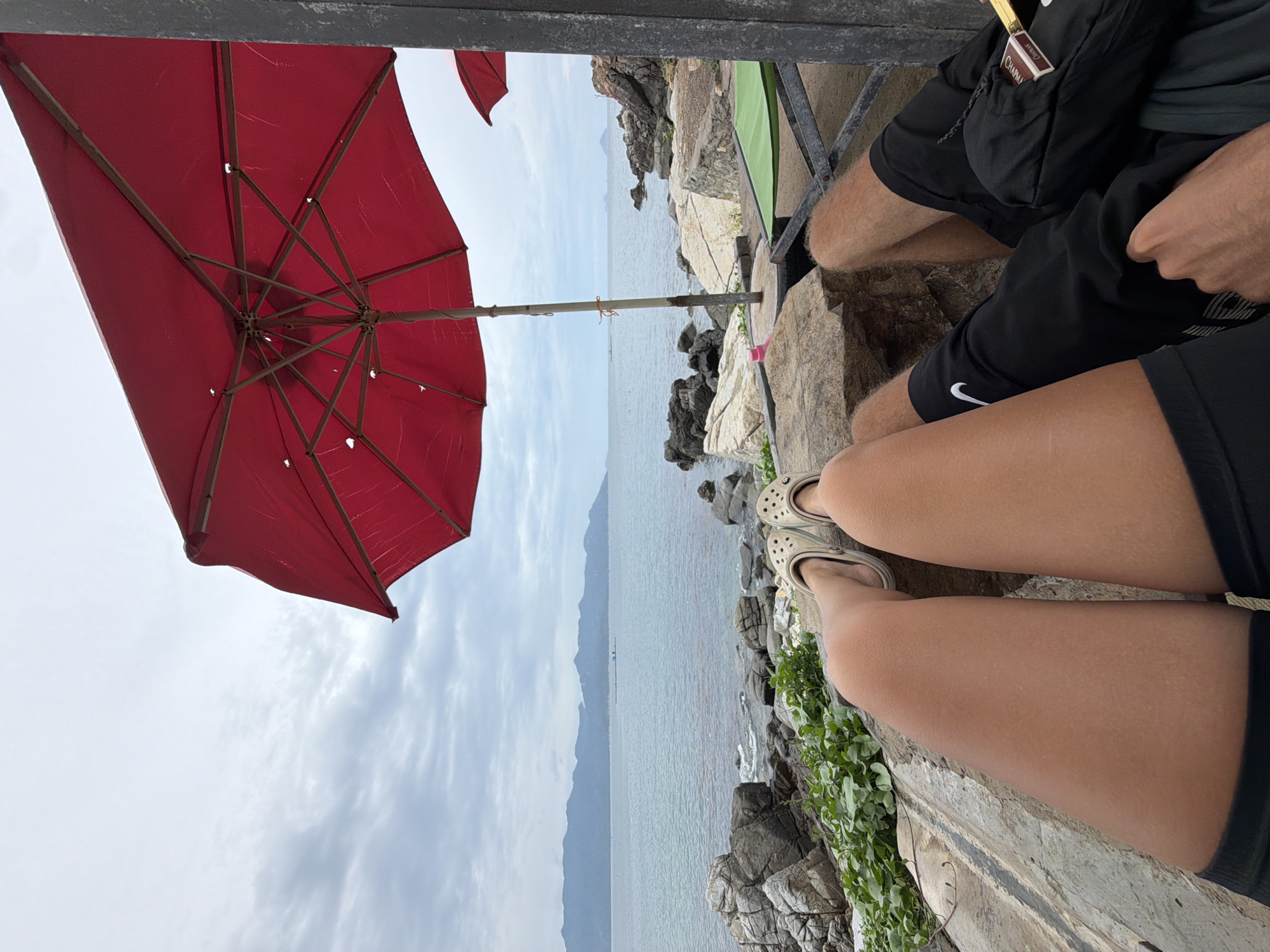 The image, originally oriented vertically, captures a relaxed coastal scene in Vĩnh Lương, Vietnam, from a seated perspective. The sky is overcast with soft clouds, suggesting a mild or cloudy day.
In the foreground, the lower bodies of at least two individuals are visible. One person wears black shorts with a white Nike swoosh logo and beige Crocs-style shoes, resting their legs on a concrete or stone ledge. Another person, to their right, also wears black shorts, with a small rectangular object, bearing the text "CHAPMAN," tucked into a pocket. Green foliage grows along the rocky structure beneath them.
A large, open red umbrella, providing shade, dominates the left side of the frame. Its internal frame and fabric structure are clearly visible from underneath. The umbrella is positioned overlooking a rocky shoreline.
Beyond the immediate foreground, the calm, greyish-blue sea extends to the horizon, meeting distant, muted mountain ranges or landmasses under the cloudy sky. The coastline is rugged and rocky, with a small, partially visible green tent or tarp nestled among the rocks in the midground. The overall scene conveys a sense of peaceful relaxation and leisure by the sea.