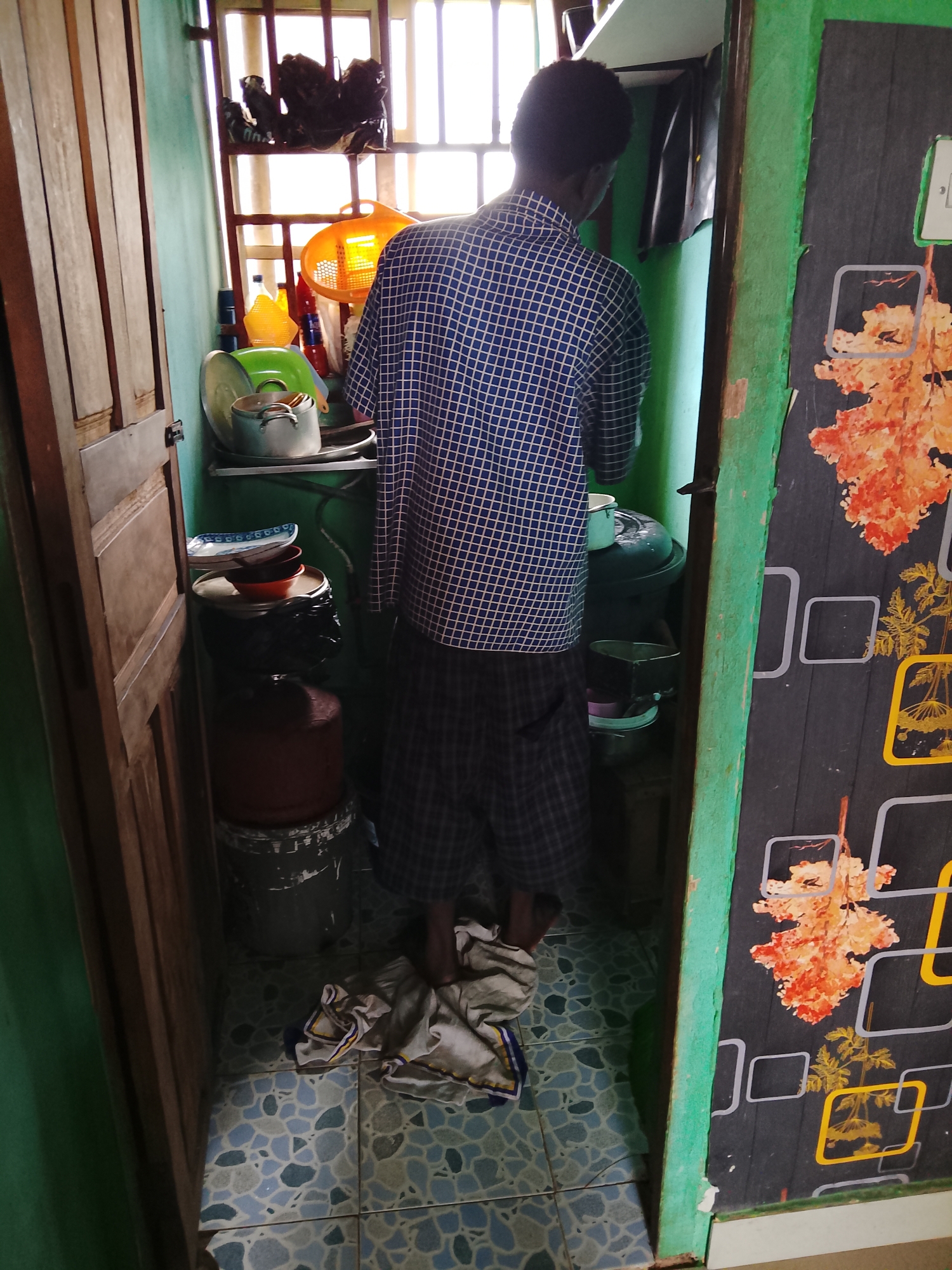 A person is standing in a small, cluttered room, facing away from the camera. The room appears to be a kitchen or pantry, with shelves filled with various kitchenware, including pots, pans, and bowls. There are also large containers, possibly for water or other liquids, and what looks like a gas cylinder. The walls are painted green, and the floor is tiled with a mosaic pattern. A wooden door is visible on the left, and a decorated panel, possibly a cabinet door, is on the right. The person is wearing a blue and white checkered shirt and plaid shorts. A crumpled cloth lies on the floor near their feet. The scene is lit by natural light coming from a window with a slatted covering at the back of the room, suggesting it is daytime. The overall impression is one of a functional, lived-in space, likely in a home in Ihiagwa, Nigeria.