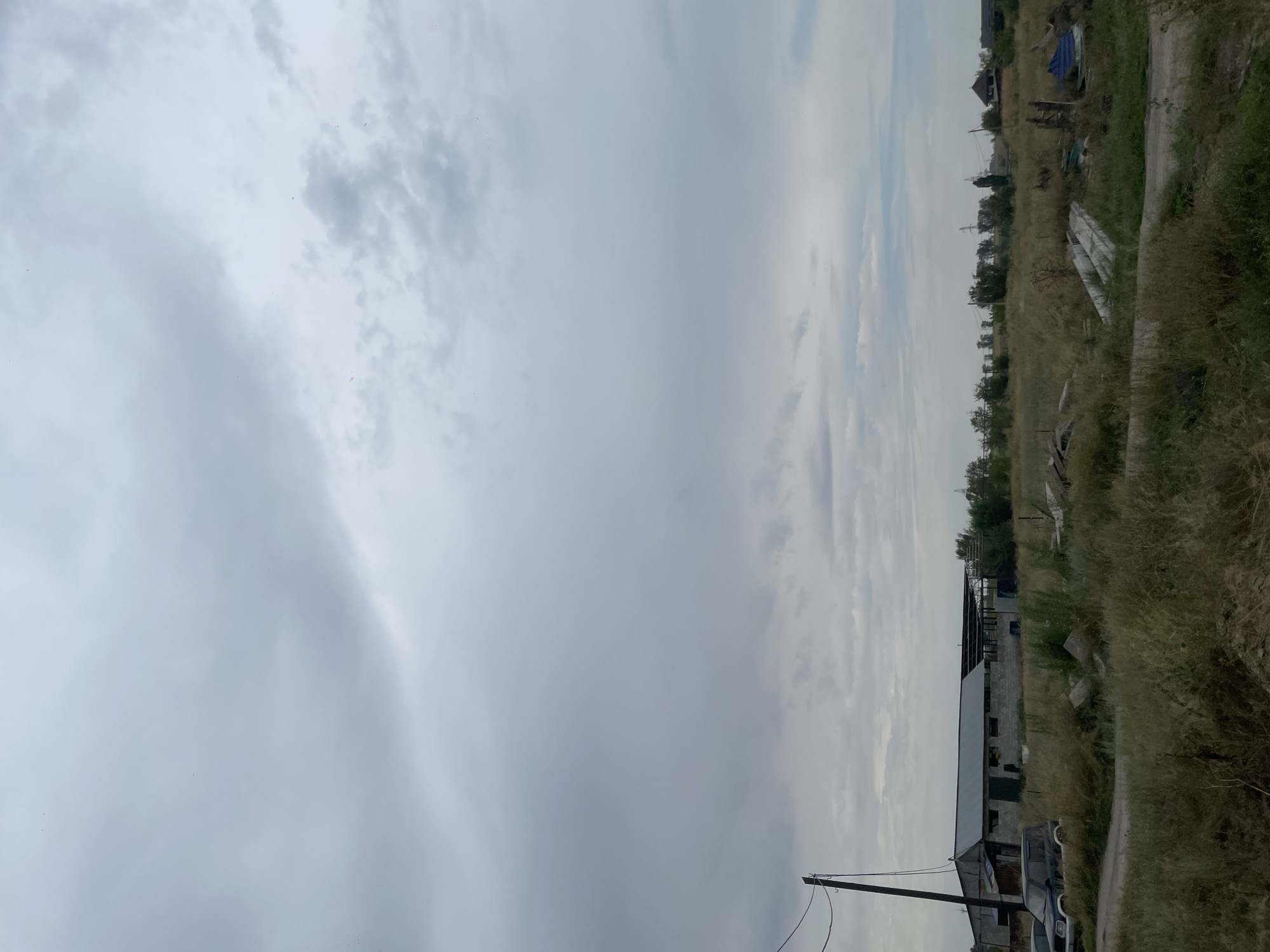 The image depicts an overcast sky over a rural landscape in Kénen Ázіrbayev aýyly, Kazakhstan. In the foreground, dry, tall grass covers the ground, with a dirt road winding through it. A few scattered concrete slabs and debris are visible in the grass. In the midground, a series of low-lying buildings with corrugated metal roofs are present, appearing to be agricultural or storage structures. A dark-colored SUV is parked to the left of the buildings. A wooden utility pole with wires stretches up towards the sky on the far left. The background shows a line of trees and distant structures, including what appear to be power lines and a residential house with a dark roof. The overall impression is one of a quiet, somewhat unkempt rural setting under a heavy, cloudy sky.