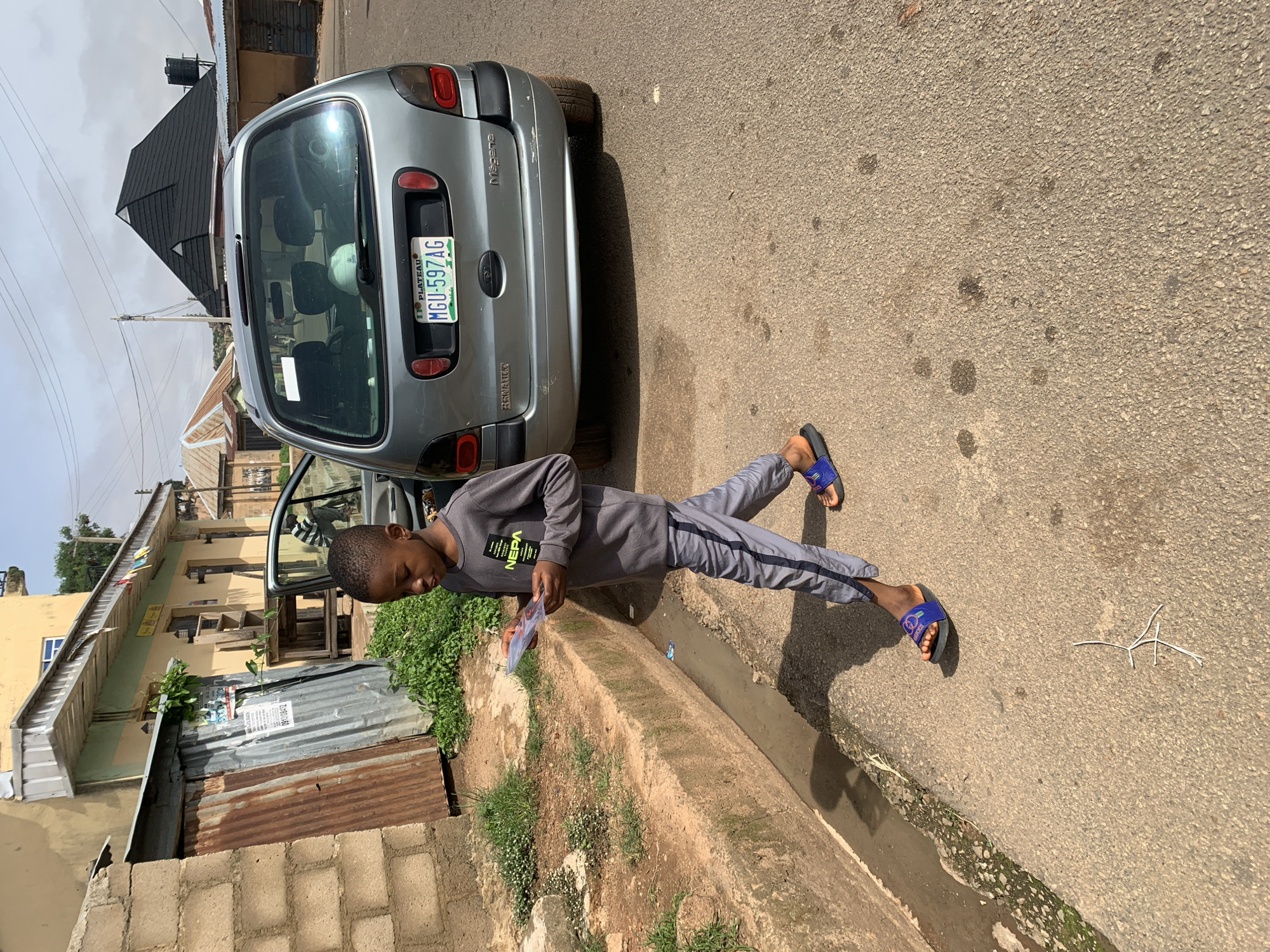 A young Black boy, wearing a gray long-sleeved shirt with "NEPA" text and gray sweatpants with a dark stripe, walks on a paved road. He is holding a thin, rectangular object, possibly a pamphlet or a thin book, in his hands and looking down at it. He is wearing blue flip-flops with a black sole. The boy is walking in front of a silver Renault Megane hatchback. The car's driver-side door is open, and its license plate reads "MGU-597AG PLATEAU". The scene appears to be in an urban or semi-urban environment in Jos, Nigeria, characterized by its architecture and road conditions. In the background, there are buildings with corrugated iron roofs and a prominent black, pyramid-shaped structure. A drainage ditch runs alongside the road. The sky is overcast with some visible utility poles and wires.