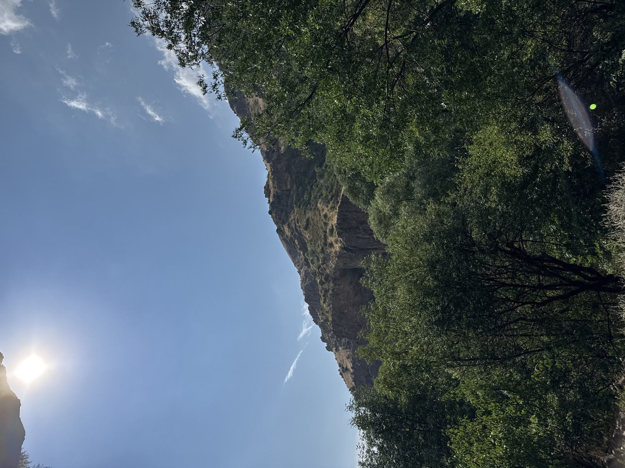 The image shows a scenic view of a mountainous area in Goght, Armenia. The foreground is dominated by lush green trees, with dense foliage. In the background, a rugged, rocky mountain slope rises against a clear blue sky. The sun is visible in the upper left corner, casting a bright glare. There are some scattered white clouds in the sky and a faint contrail. The overall impression is one of natural beauty and tranquility.