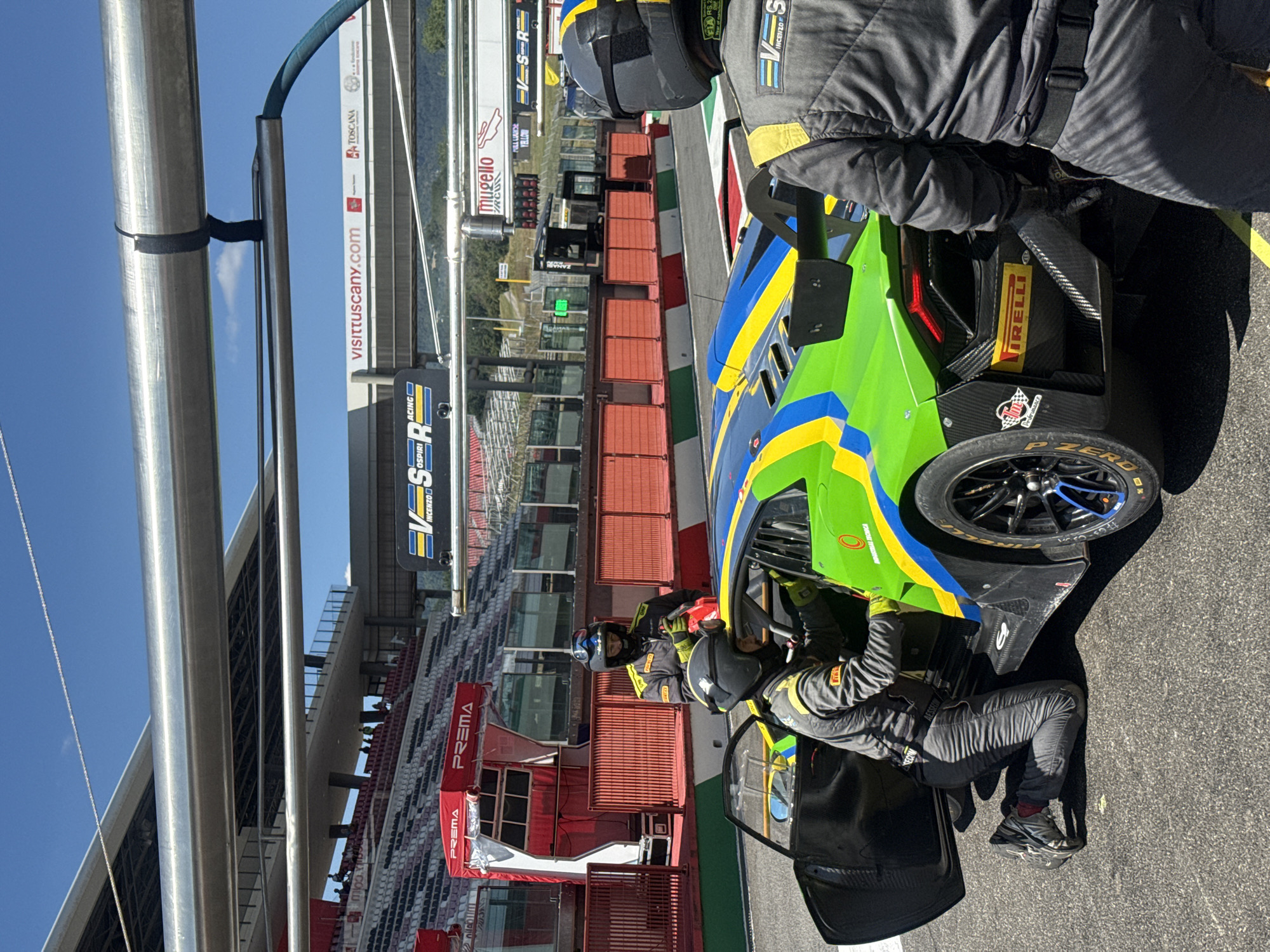 In Scarperia, Italy, at the Mugello Circuit, a pit stop is underway for a green and blue race car, likely a Lamborghini Huracan GT3 based on its distinctive shape and livery. Two individuals in racing suits and helmets are attending to the car. One person is kneeling beside the open driver's side door, appearing to be working on the interior or adjusting something within the cockpit. The second person is standing slightly behind the kneeling individual, holding a red object, possibly a tool or equipment, and wearing yellow gloves. A third person, also in a racing suit and helmet, stands to the right of the frame, facing away from the camera, providing a rear view of their attire. The background shows the grandstands of the racetrack, a red tent structure with "PREMA" branding, and various signage, including "VISIT TUSCANY.COM" and "VS RACING". The scene is illuminated by bright sunlight, suggesting it is daytime.