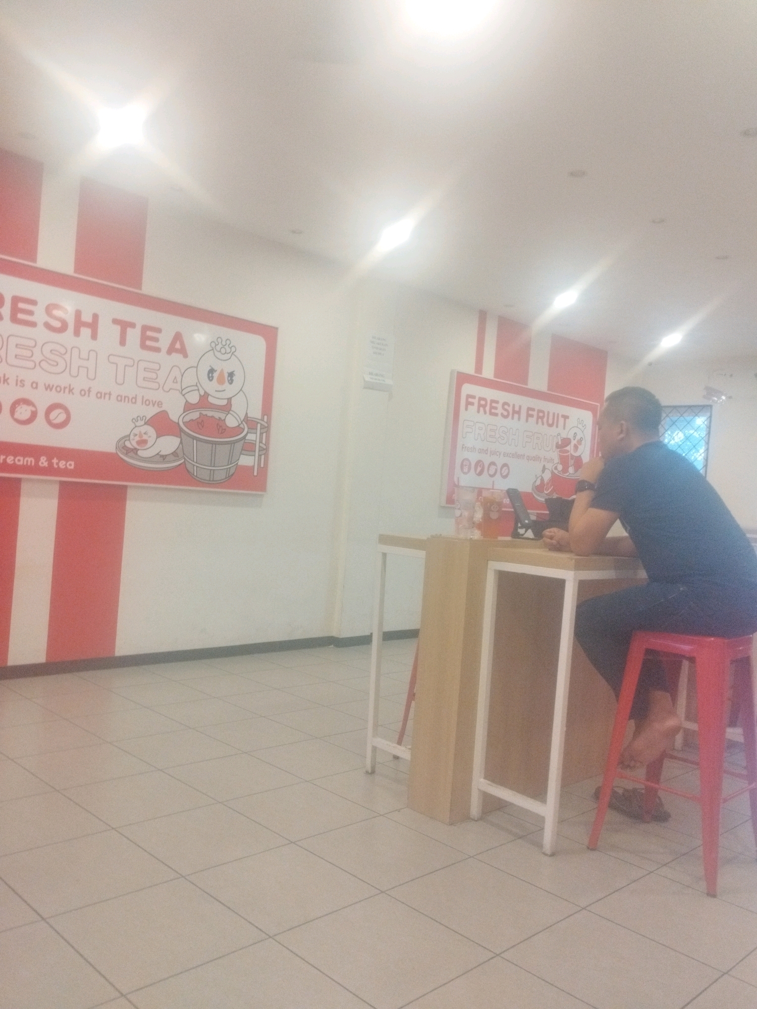 The image shows the interior of a "Fresh Tea" shop, likely located in Percut Sei Tuan, Indonesia, given the context. A man is seated at a counter, facing away from the camera and towards a tablet device on the counter. He is wearing a dark blue t-shirt and dark pants, and is sitting on a red stool. The counter is made of light-colored wood with white metal legs. On the wall behind the counter are two signs. The sign on the left has pink and white stripes and displays cartoon characters and text related to "Fresh Tea." The sign on the right also has pink and white stripes and promotes "Fresh Fruit" with similar cartoon imagery and text. The floor is tiled in a light beige color. The lighting appears to be from overhead fluorescent lights, with some glare visible.