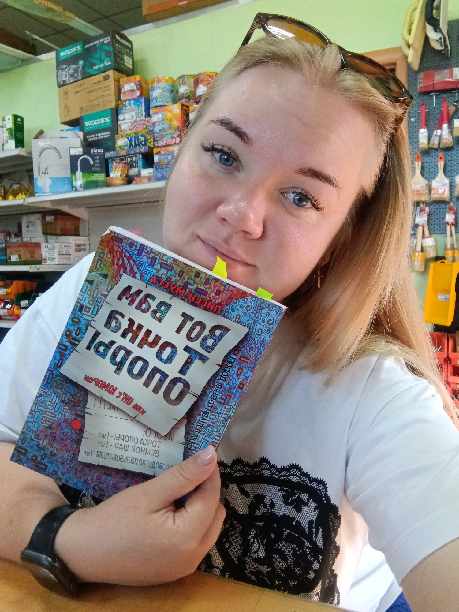 A fair-skinned woman with blonde hair and blue eyes is holding a book with a blue and red patterned cover in front of her face. She is wearing a white t-shirt with a black lace design on the front. Sunglasses are perched on top of her head. The background appears to be a store aisle, with shelves stocked with various items, including boxes, paintbrushes, and other hardware supplies. The overall lighting suggests an indoor environment with artificial light.