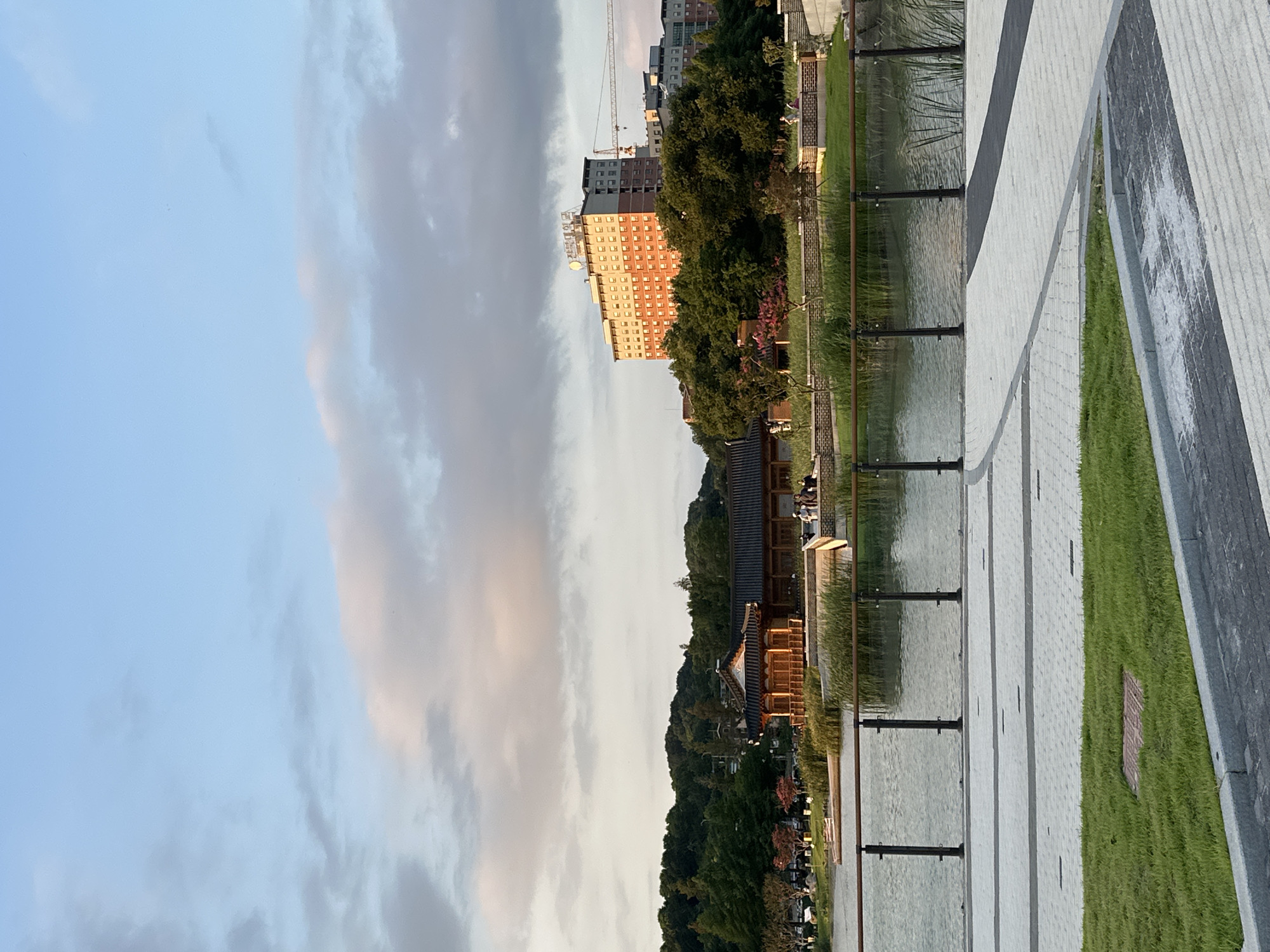 The image depicts a serene outdoor scene at dusk in Jeonju-si, South Korea, likely a public park or waterfront area. In the foreground, a paved walkway with distinct patterns of light and dark gray rectangular stones leads to a neatly manicured grassy area bordered by a concrete curb. A dark railing with glass panels separates the walkway from the water's edge.
Beyond the railing is a tranquil body of water, reflecting the soft hues of the sky. Tall reeds and grasses grow along the near bank of the water. In the middle ground, a traditional Korean-style wooden pavilion with a dark, curved roof stands prominently. Several figures are visible near the pavilion, appearing to be people walking or conversing. Behind the pavilion, a lush green hillside is covered with dense trees.
Dominating the background is a large, multi-story building with a light-colored facade and orange-tinted windows, resembling an apartment complex or hotel. The building is topped with a sign displaying Korean characters. To the right of this building, a construction crane is visible, suggesting ongoing development in the area. The sky above is a soft blue, with wispy clouds tinged with pink and orange from the setting sun. The overall atmosphere is peaceful and picturesque.