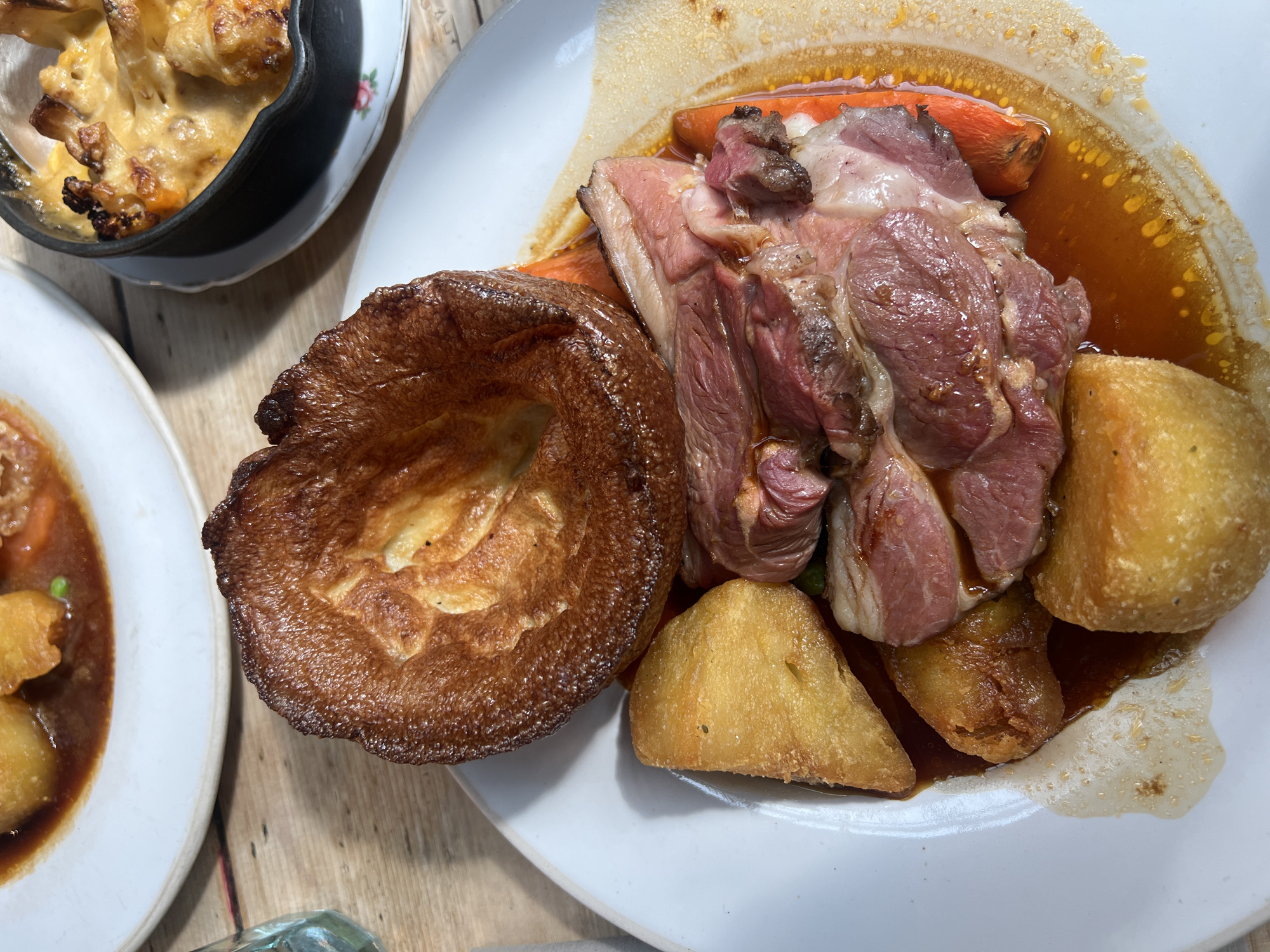 The image shows a roast dinner meal on a white plate, likely taken in London, United Kingdom. The main components of the meal are Yorkshire pudding, roasted meat (possibly lamb), roasted potatoes, and sliced carrots, all covered in gravy. A second plate to the left has more roasted potatoes with gravy. A small black pot containing what appears to be cauliflower cheese is present on a white plate at the top left of the image. The scene is on a wooden table.