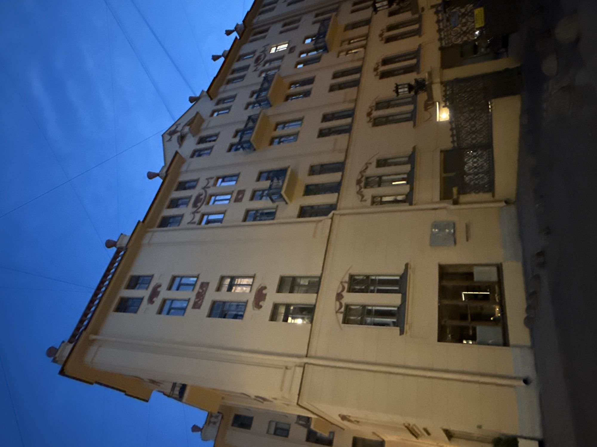 The image shows a multi-story building with a light-colored facade against a dark blue sky, suggesting it is twilight or early evening. The building has numerous windows, some of which are illuminated from within, indicating occupancy. Decorative elements are visible on the facade, including what appear to be stylized animal motifs or crests above some of the windows, and decorative patterns above others. The building appears to be in an urban setting, as evidenced by the presence of overhead wires and the dark, shadowy area to the right which could be a street or another building. The architecture suggests a historical European style, consistent with a location like Saint Petersburg, Russia. There is no clear indication of any specific activity or event taking place, other than the general presence of life within the building as suggested by the lit windows. No text is visible in the image. The overall mood is somewhat subdued, fitting for the late hour and urban environment.