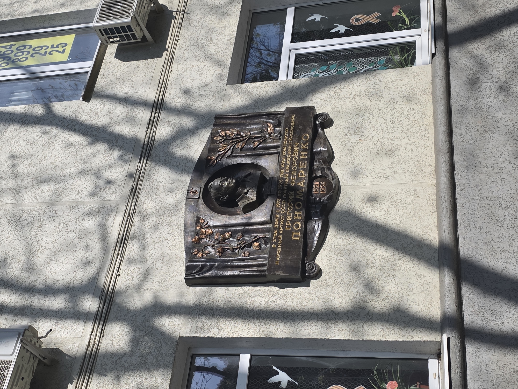 The image captures a bronze memorial plaque affixed to a building in Krasnodar, Russia, commemorating Grigory Fedorovich Ponomarenko, a composer and People's Artist of the USSR. The plaque features a relief portrait of Ponomarenko along with a stylized inscription in Russian. The text indicates that he lived in this building from 1973 to 1996 and was an honorary citizen of Krasnodar. The building's facade is light-colored with a rough textured surface. Two windows are visible above and below the plaque. The top window has white frames and decorative paper cut-outs of birds and a ribbon, with a plant visible inside. The lower window has similar frames and a single cut-out of a bird. An air conditioning unit is located to the left of the plaque, along with visible conduit running down the side of the building. Shadows of tree branches are cast across the wall.