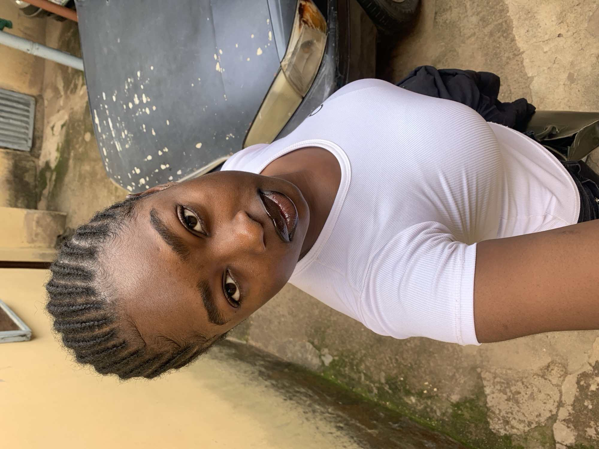A young Black woman with braided hair is taking a selfie outdoors. She is wearing a white ribbed t-shirt and dark pants. She is standing next to a light-colored wall and a dark-colored vehicle with visible damage. The ground is paved with cracked concrete. The location is identified as Rafin Par, Nigeria.