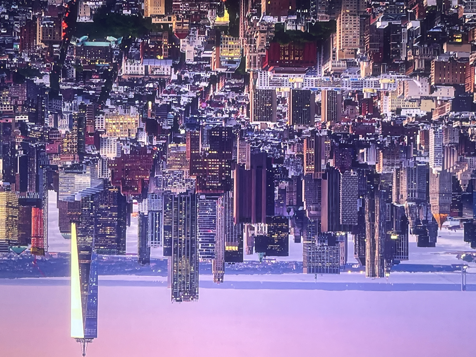 The image is an aerial view of a densely populated cityscape at dusk, with a purple and pink sky. The architecture is predominantly modern, featuring numerous skyscrapers of varying heights and designs. Lights are visible in many windows, indicating that it is nighttime or late evening. In the foreground, there are many smaller buildings, also lit up. In the background, a body of water can be seen, with a bridge and distant landmass. The most prominent structure in the image is a very tall, slender skyscraper with a distinctive illuminated spire, which appears to be One World Trade Center. The overall impression is of a bustling metropolis at the end of the day.