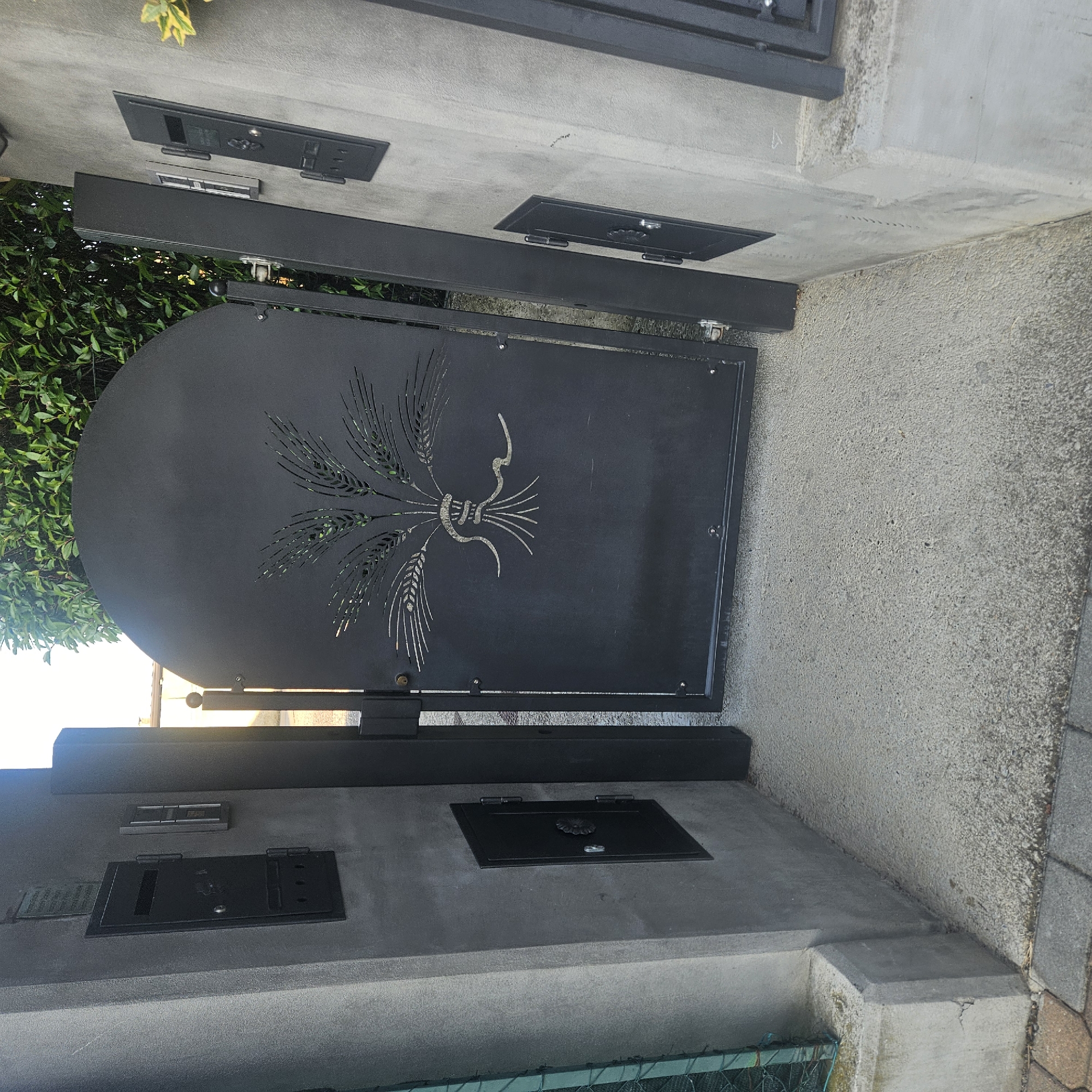 The image shows an outdoor scene in Calcinate, Italy, focusing on a dark metal gate with an arched top, featuring a laser-cut design of wheat stalks and a snake. The gate is open, revealing a glimpse of green foliage behind it. To the left of the gate is a concrete pillar with a black mailbox or parcel locker embedded in it, and below that, a smaller black recessed box with a decorative handle. To the right of the gate, another concrete pillar is visible, also with a black mailbox or parcel locker. The ground in front of the gate is paved with concrete, and to the left, there is a glimpse of a paved pathway with cobblestones. The overall impression is of a well-maintained entrance to a property.