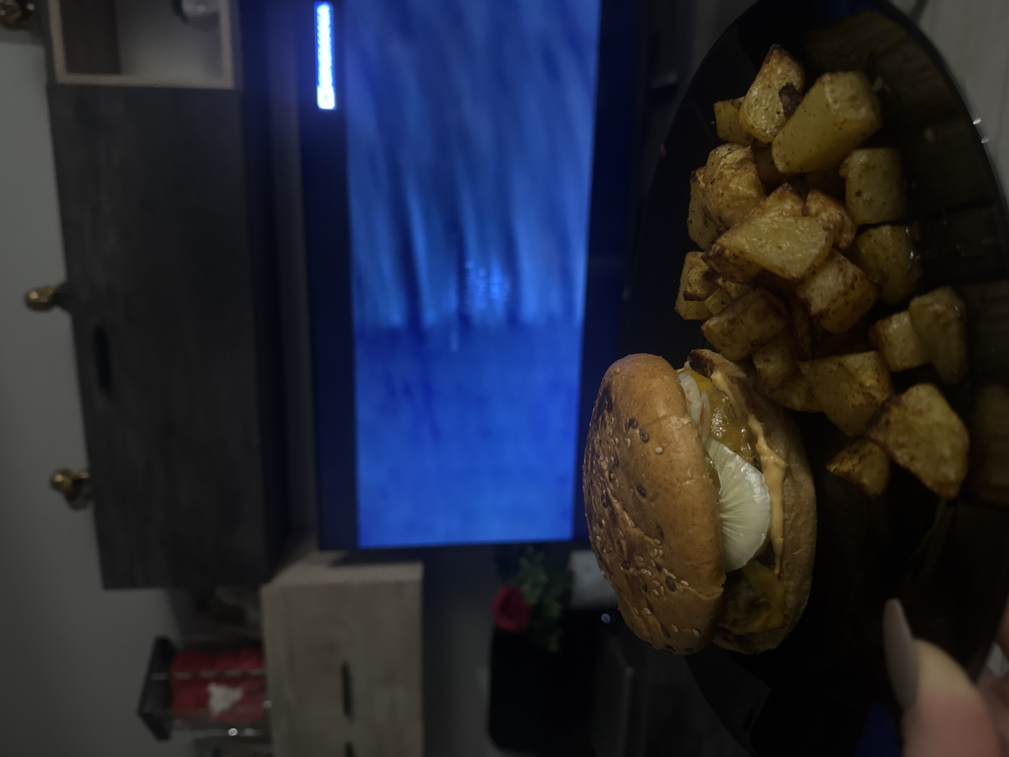 The image depicts an indoor scene, likely a living room or dining area in Varna, Bulgaria, illuminated by artificial light, suggesting evening or nighttime. The primary subject is a meal presented on a black plate, held in the bottom right by a person's hand, identifiable by a long, light-colored fingernail.
The meal consists of a well-prepared burger and a generous serving of roasted or fried potato chunks. The burger features a sesame seed bun, a patty, melted cheese, visible slices of white onion, and a creamy sauce.
In the background, a large television screen displays a predominantly blue image, which could be a video, game, or a static graphic. On the left side of the screen, vertically oriented white text is partially visible, appearing to spell "SAMSUNG," indicating the brand of the television or an interface element. Below the television, a dark-colored piece of furniture, possibly a TV stand or cabinet, is present. A small red decorative object, resembling a rose, can be seen in front of the TV stand. Further below, a small red box with a white play button triangle, indicative of the YouTube logo, is visible. The overall setting suggests a casual and relaxed atmosphere for enjoying a meal while watching television.