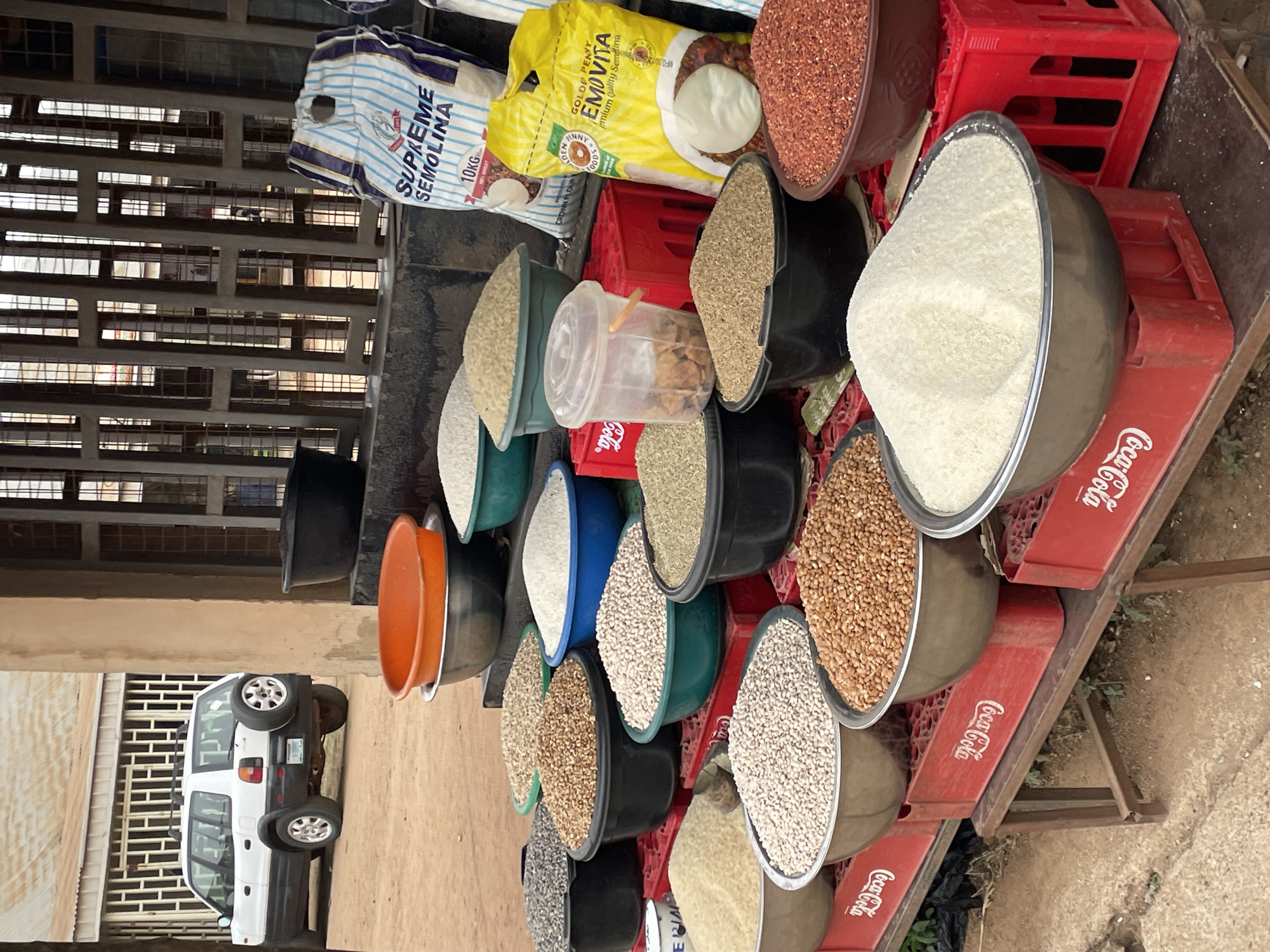 The image depicts a roadside stall selling grains and legumes, likely in Jos, Nigeria. Several containers are arranged on red Coca-Cola crates, filled with various products. There are bags of "SUPREME SEMOLINA" and "GOLDE PENNY EMOVITA," indicating common food staples. The containers are diverse in color and material. Behind the stall, a white vehicle is parked along a street with buildings and a gated window structure. The overall setting appears to be an informal marketplace.