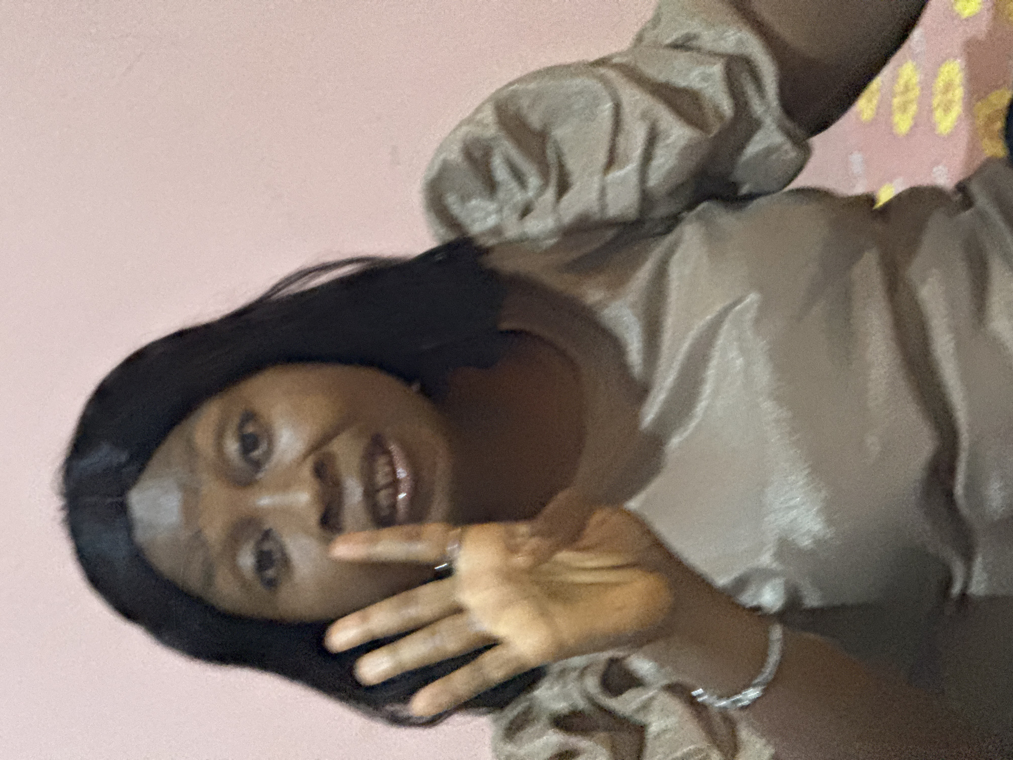 An indoor portrait features a woman with dark skin and long, dark hair, looking directly at the camera with a slight smile. She is wearing a light brown or tan top with voluminous, puffy sleeves, made of a shiny fabric. On her left wrist, she has a silver-colored bracelet, and a ring is visible on her right ring finger. Her right hand is raised with the palm open, positioned in front of her face, possibly in a wave or greeting gesture.
The background behind her head and upper body is a plain, light pink wall. To the lower right side of the frame, a small section of a pink fabric with yellow, geometric patterns can be seen, which might be upholstery or another decorative item. The image is lit from an unseen source, creating soft shadows, and has a slightly grainy texture. The overall impression is an informal, candid moment. There is no visible text in the image.