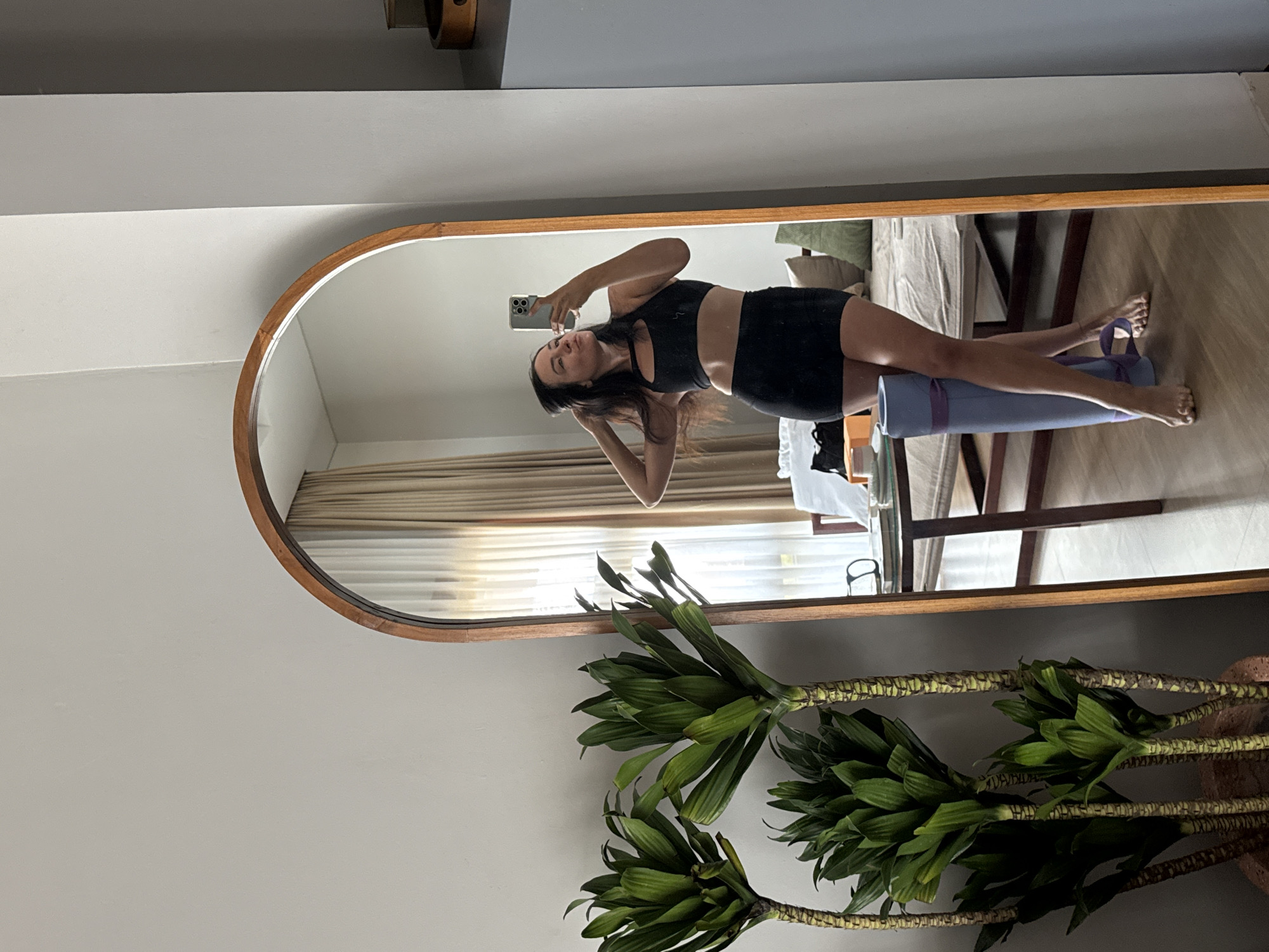 A woman stands in front of a tall, arched mirror, taking a selfie. She is wearing a black sports bra and a black skirt. Her hair is dark and wavy, and she has her hands raised to her head. In the reflection, a yoga mat is visible leaning against her legs. The background shows a room with a couch, a table, and curtains. Two tall potted plants with green leaves are in the foreground, to the left of the mirror. The environment suggests a home or a yoga studio. The location is specified as North Kuta, Indonesia.
