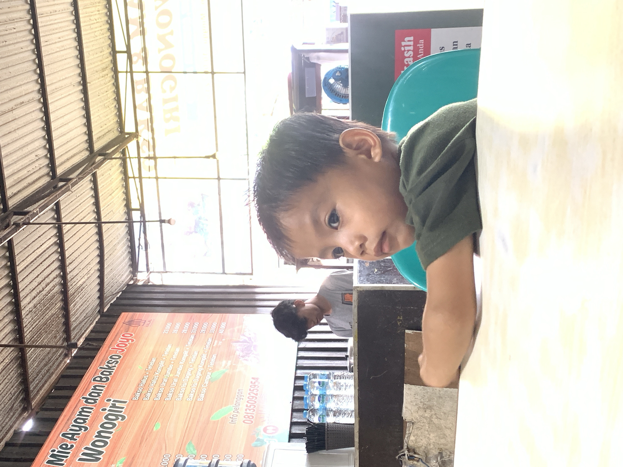 A child sits at a table in an outdoor eatery. The eatery is called "Mie Ayam dan Bakso Joyo Wonogiri", and the sign lists various "Bakso" dishes and prices in Indonesian Rupiah. The menu includes "Bakso Halus + Tetelan", "Bakso Halus Komplit + Tetelan", "Bakso Urat Jumbo + Tetelan", "Bakso Urat Jumbo Komplit + Tetelan", "Bakso Isi Daging + Tetelan", and "Bakso Campur + Tetelan." A man in a grey shirt sits at a table nearby. Bottles of drinking water are on the table. The eatery is located in Samarinda, Indonesia, as indicated by the phone number with Indonesian country code on the menu.
