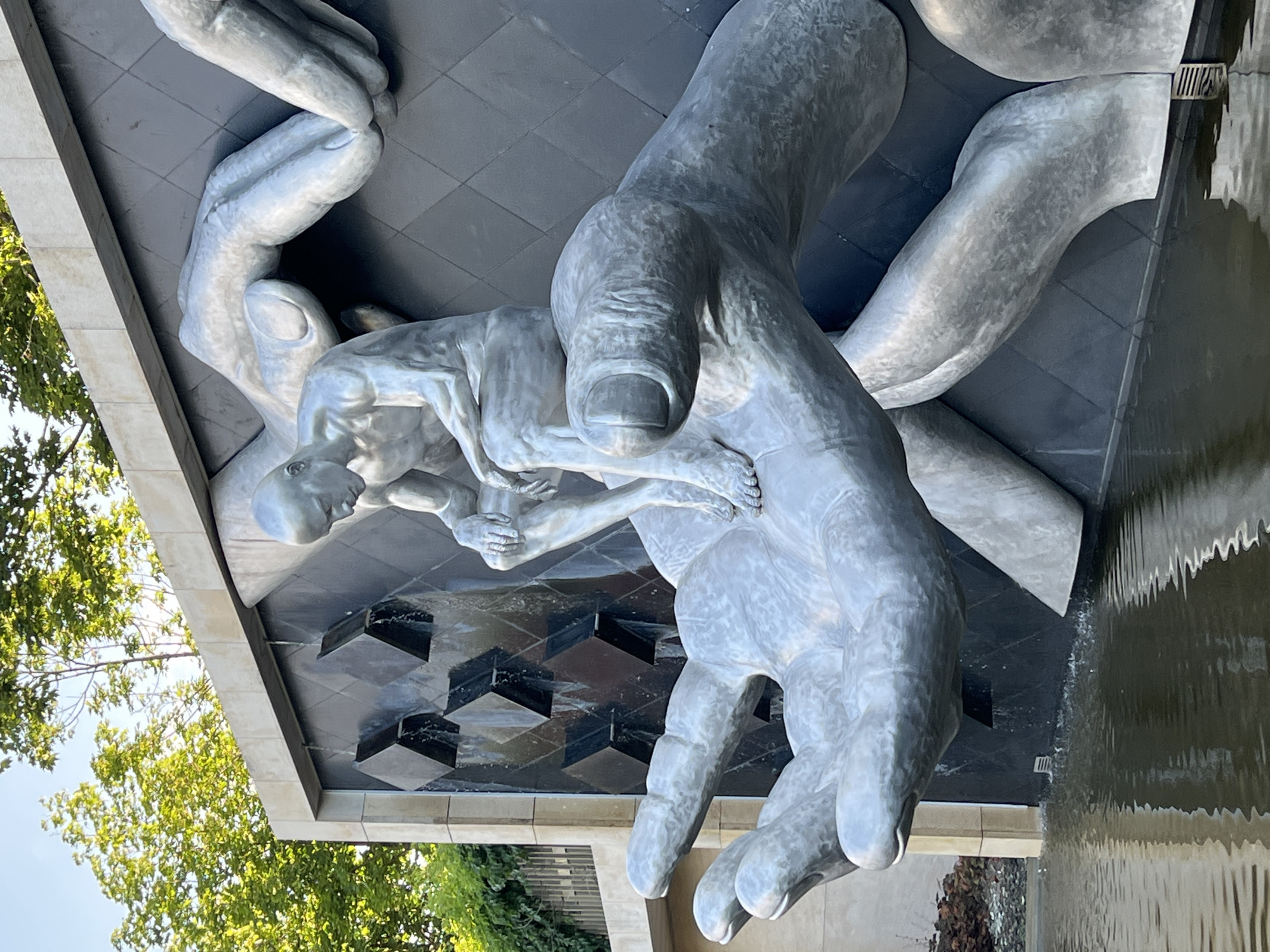 The image shows a sculpture of a seated man on a backdrop of a dark, textured wall with water cascading down. The man is depicted in a contemplative pose, with his head bowed and his hands resting on his knees. He appears to be made of a metallic material, with a polished, silver finish. Surrounding the seated figure are several large, abstract hands, also made of the same metallic material, which seem to be cradling or supporting the man. The sculpture is situated in a setting with lush green trees and a body of water in the foreground, reflecting the sculpture and the surrounding environment. The water is calm, with ripples on the surface. The background features architectural elements, including a light-colored stone structure and a darker, textured wall with geometric patterns. The overall scene suggests a public art installation, likely in an outdoor urban space. This sculpture is located in Krasnodar, Russia.