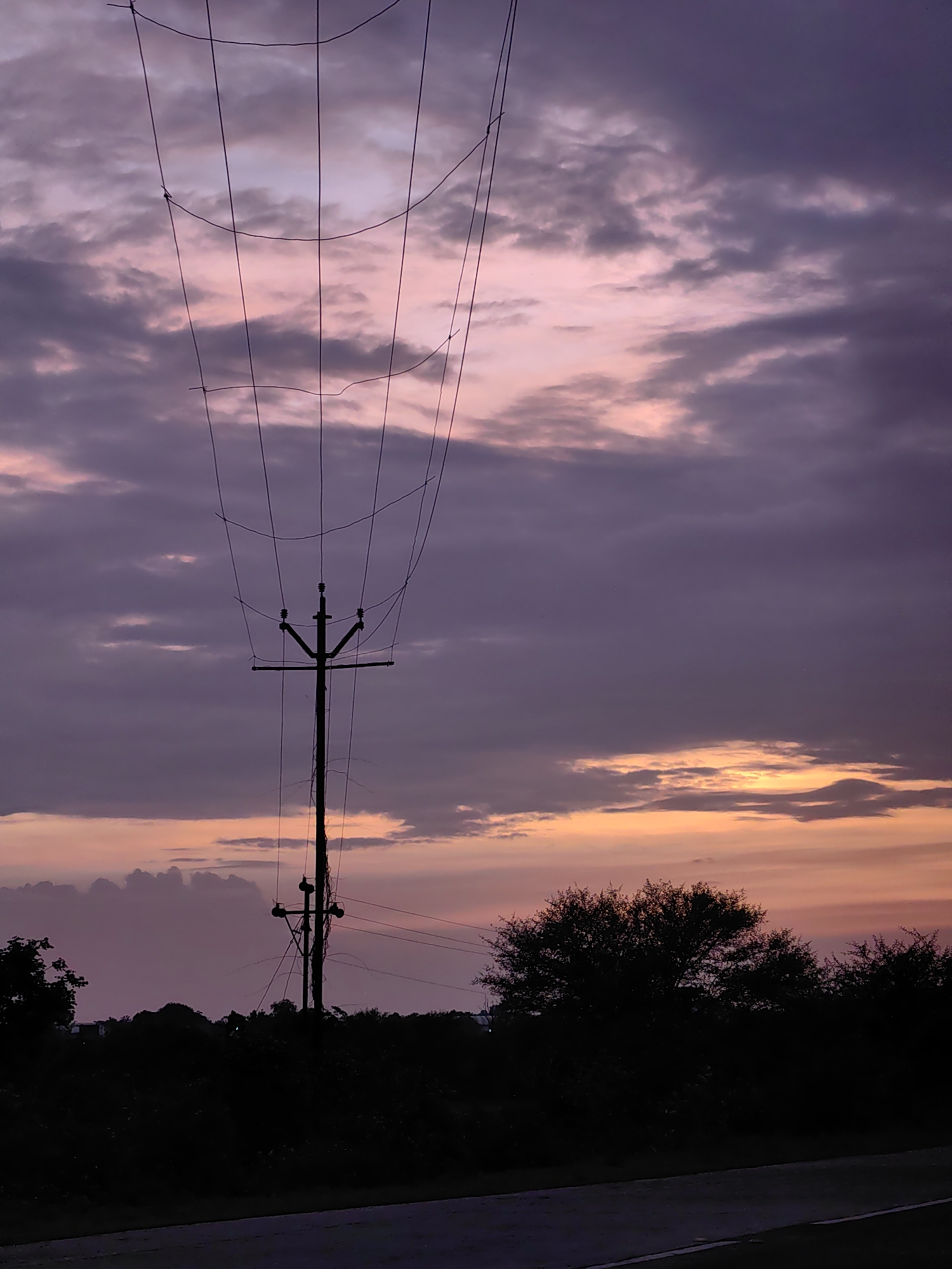 This image captures a serene sunset scene in Dattapur, India. The sky is a canvas of soft purples and pinks, with streaks of orange peeking through the clouds near the horizon. A solitary utility pole stands silhouetted against the vibrant sky, its wires stretching across the frame. Below, a dense line of dark trees forms the horizon, with a paved road or path in the foreground. The overall impression is one of peaceful twilight in a rural or semi-rural setting.