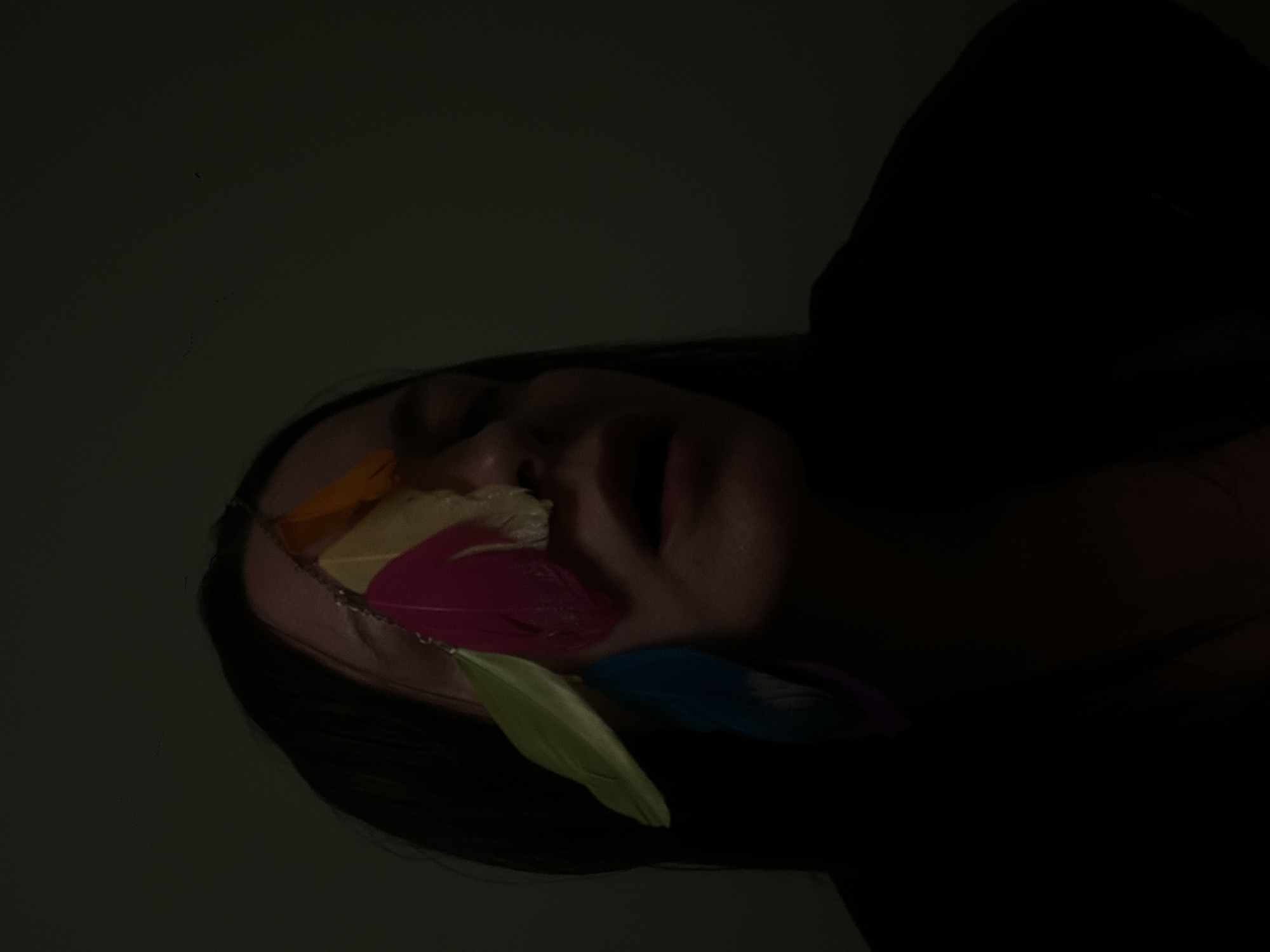 The image presents a close-up of a person's face, partially obscured by darkness, with a headdress featuring colorful feathers (orange, yellow, pink, green, and blue). The person's eyes are closed, and their mouth is slightly open. The dark background and the angle of the shot create an intimate, possibly artistic, atmosphere. No other details allow for an inference of location in Riga, Latvia.