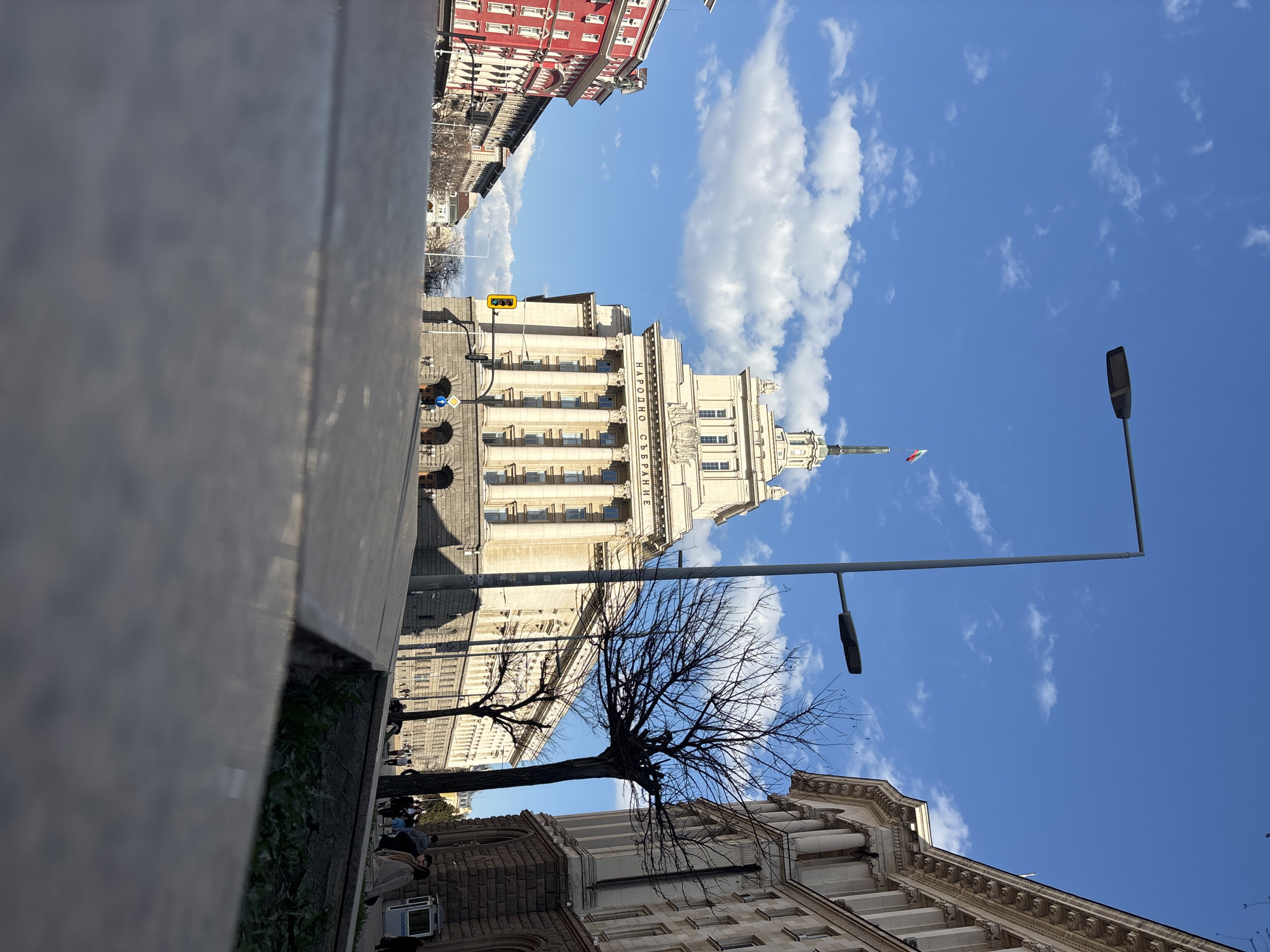 The image shows the National Assembly of Bulgaria building in Sofia, Bulgaria. The building is a large, light-colored structure with classical architectural elements like columns and a central dome. The facade bears the Cyrillic inscription "НАРОДНО СЪБРАНИЕ" (Narodno Sabranie, meaning National Assembly). To the left, a blurry stone structure is in the foreground. A bare tree is visible between two buildings. Streetlights are present. The sky is blue with scattered clouds. Buildings with red facades are visible in the background. People are walking around the building.