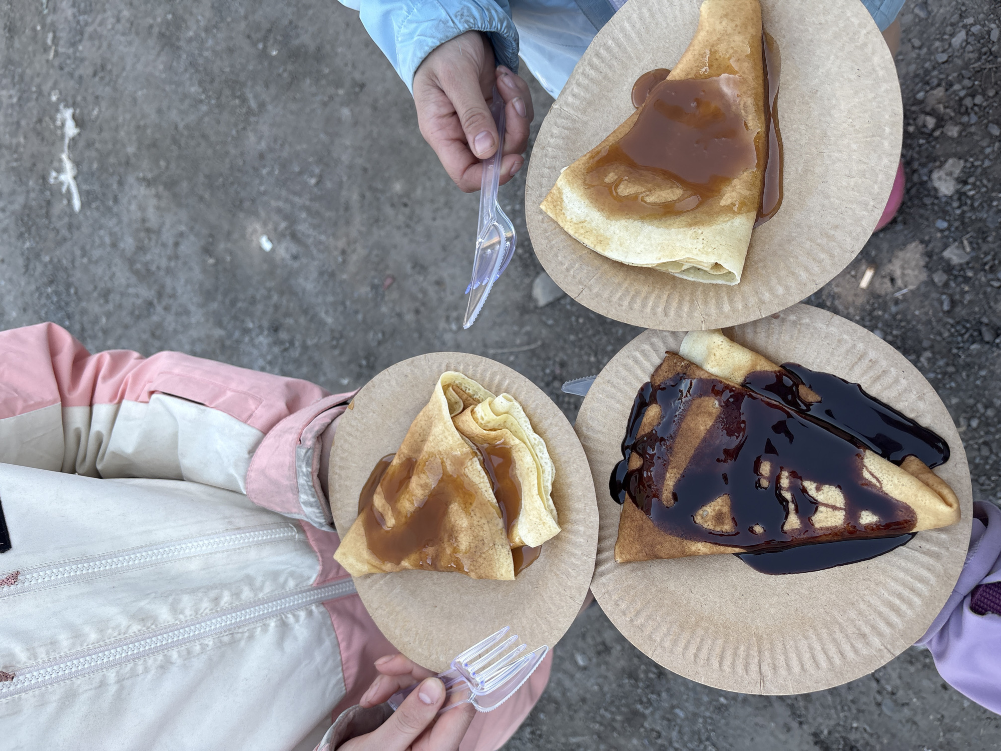 Three individuals stand on a grey, possibly asphalt or concrete, surface. Each person is holding a paper plate with a crêpe drizzled with sauce. The plate held by the person on the top right has a light brown sauce, while the plate on the bottom left has a similar light brown sauce with a smaller portion. The plate on the bottom right holds a crêpe with a dark, glossy sauce. Each person also holds a clear, plastic fork or spork. The individuals are wearing winter jackets; the person on the top right has a light blue jacket, the person on the bottom left has a pink and white jacket, and the person on the bottom right has a purple jacket. The location is unspecified, but provided information indicates the city of Nizhnie Yaki, Russia.