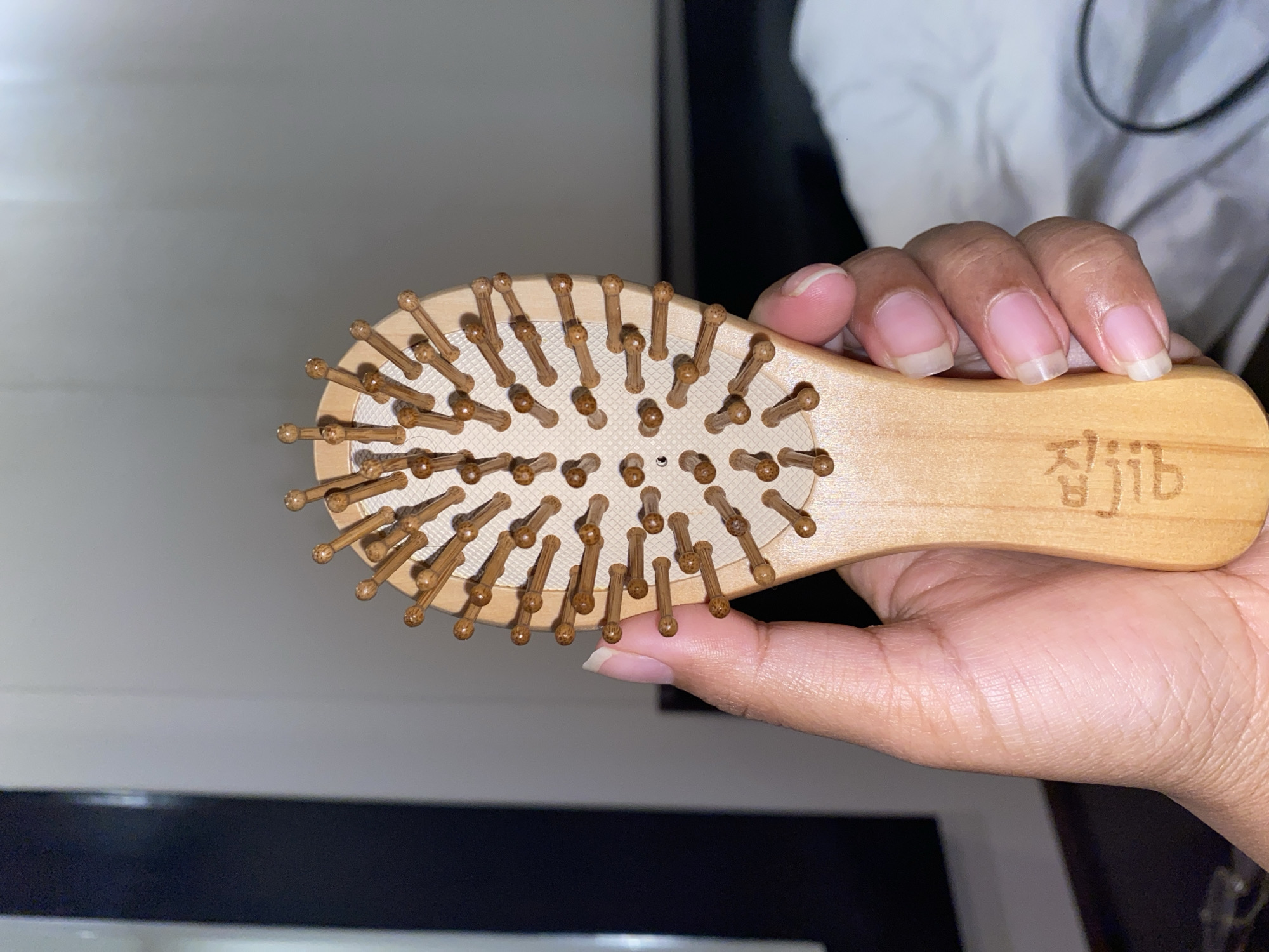 A person is holding a small, light-wood hairbrush with rounded, brown wooden bristles. The brush has a wooden handle with what appears to be Korean characters etched onto it. The person's hand has light brown skin with clean, short fingernails. The background is blurry and shows a mix of black and white surfaces. The scene is indoors. There is no indication in the image that the location is North Kuta, Indonesia.