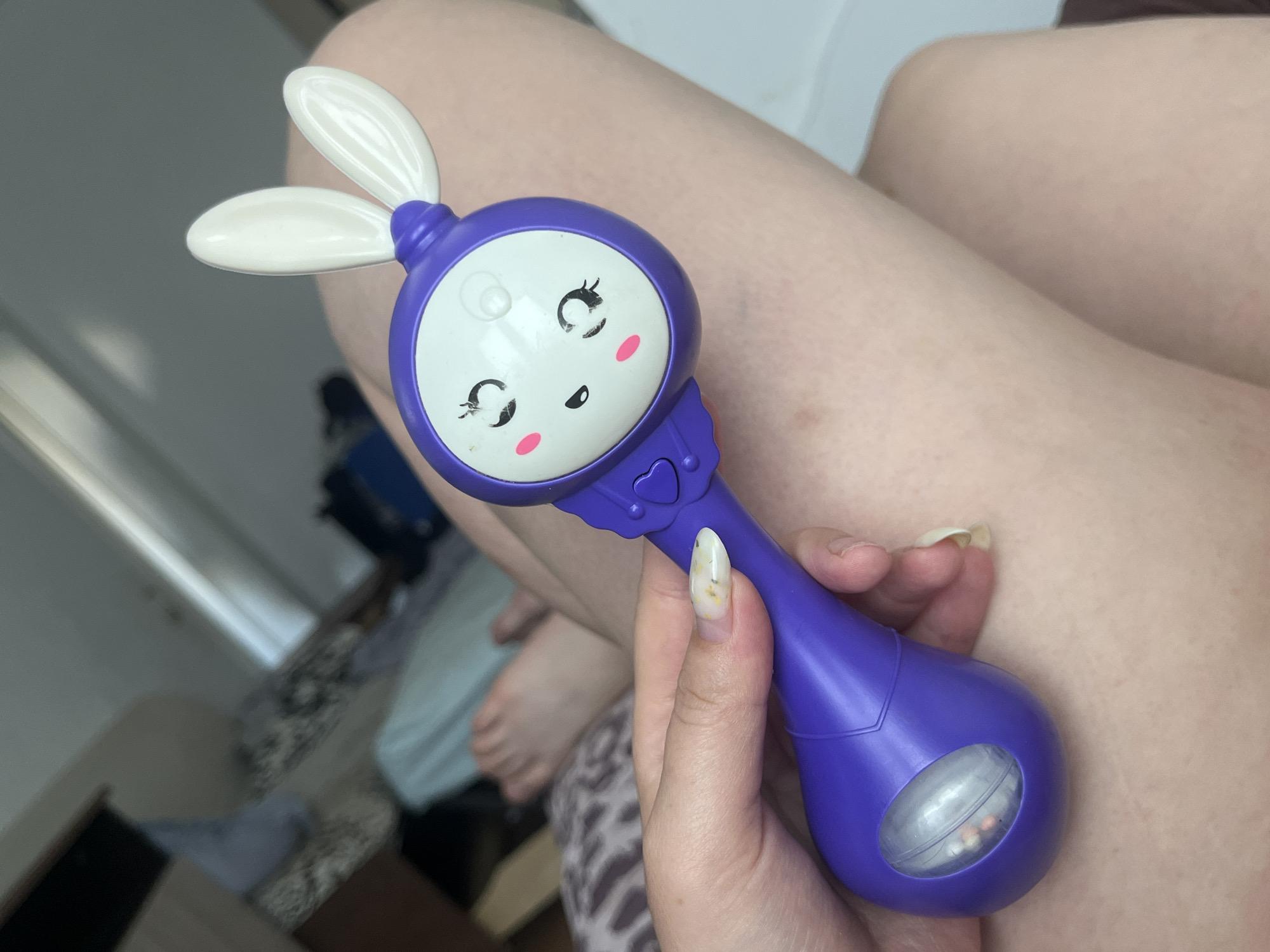 A person's hand is holding a purple toy shaped like a cartoon character with bunny ears. The toy has a white face with drawn-on eyes, nose, and rosy cheeks. The person's fingernails have a light-colored polish with small flecks of yellow and green. In the background, there is a bed with patterned bedding, and part of another person's leg is visible. The scene appears to be indoors, possibly in a bedroom. There is no information in the image to indicate the location in the city of Chelyabinsk, Russia.