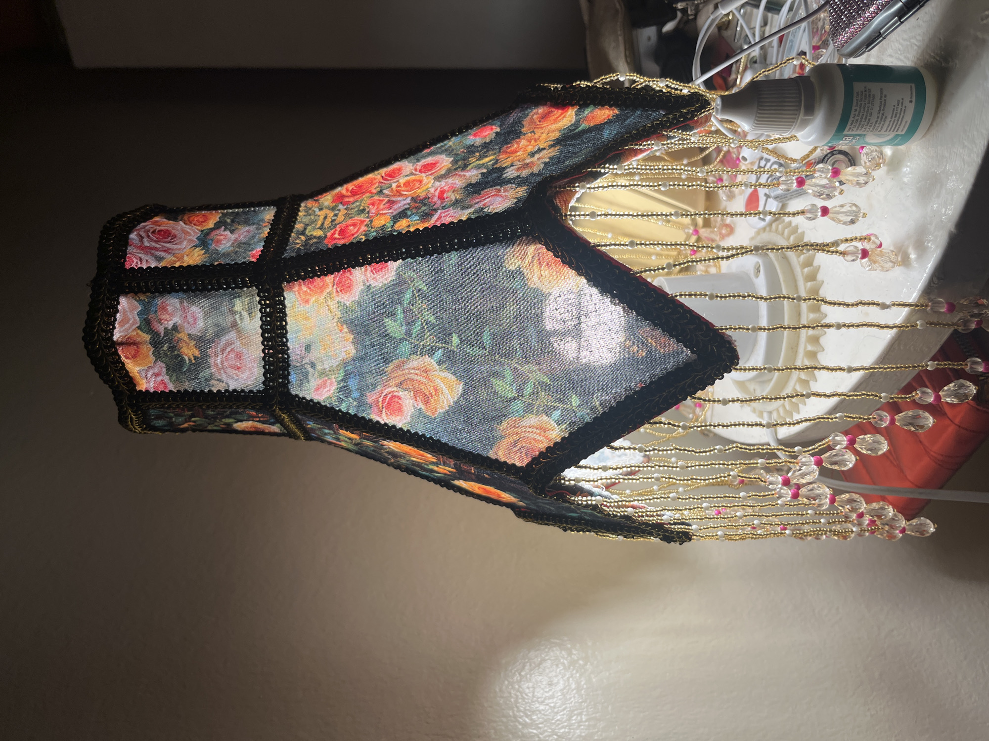 A fabric lampshade with a rose pattern and beaded trim is positioned near the edge of a white surface, possibly a table or counter. The lampshade is made of several panels connected by black trim. Beaded strings hang from the bottom of the shade. In the background, there are other objects, including wires and a bottle. The context of the location in Buenos Aires, Argentina, is not provided in the image, so the image context of Buenos Aires location is not available.