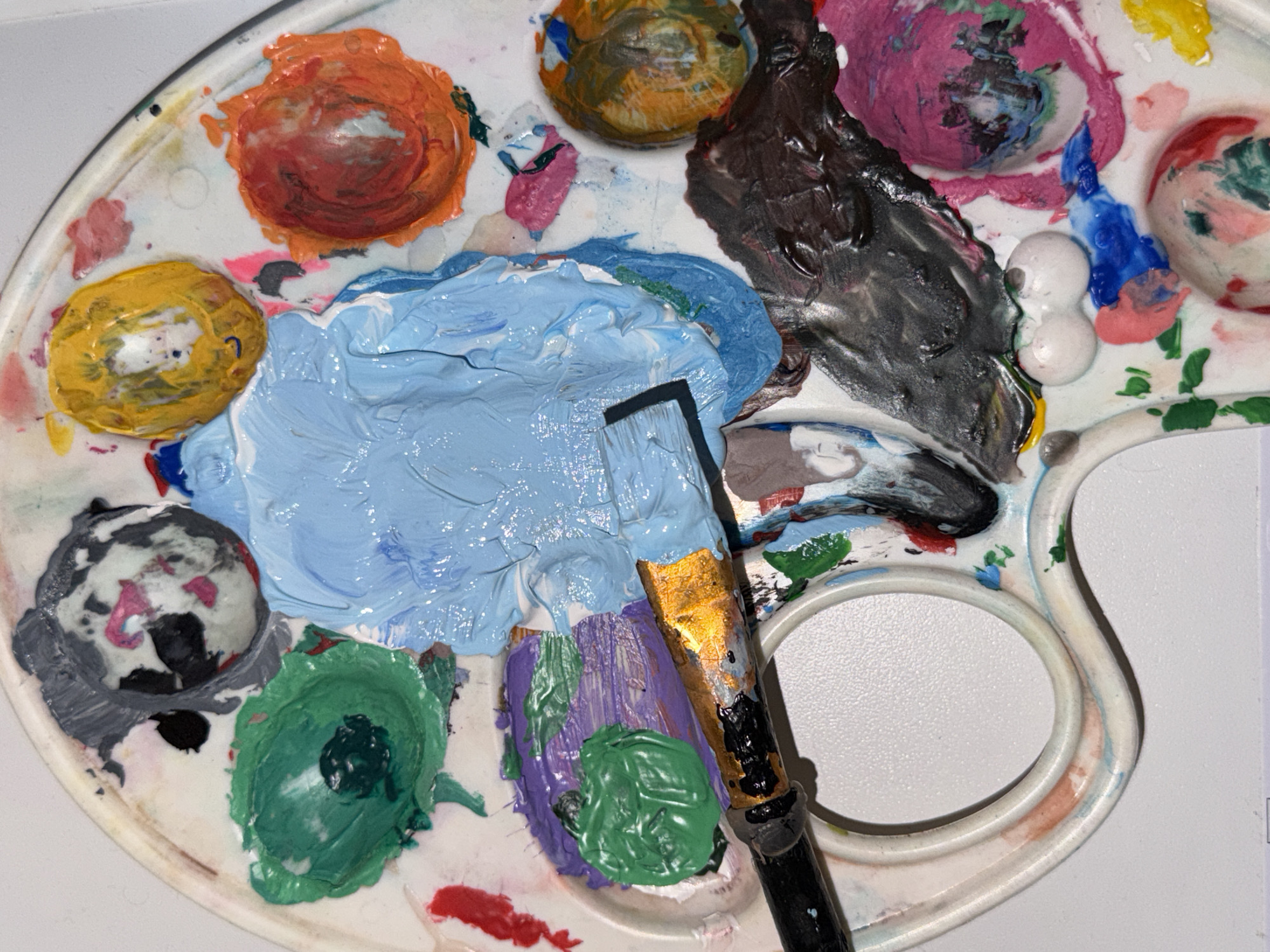A top-down view depicts a white, oval artist's palette containing various dollops of paint. The palette is filled with blobs of orange, yellow, blue, black, pink, and green paint. A paintbrush with a black handle and gold-colored bristles rests on the palette, partially covered with the same blue and purple paint. The palette's white surface exhibits further remnants of paint mixing. The background is plain and white. This scene is not attributable to Perm, Russia.