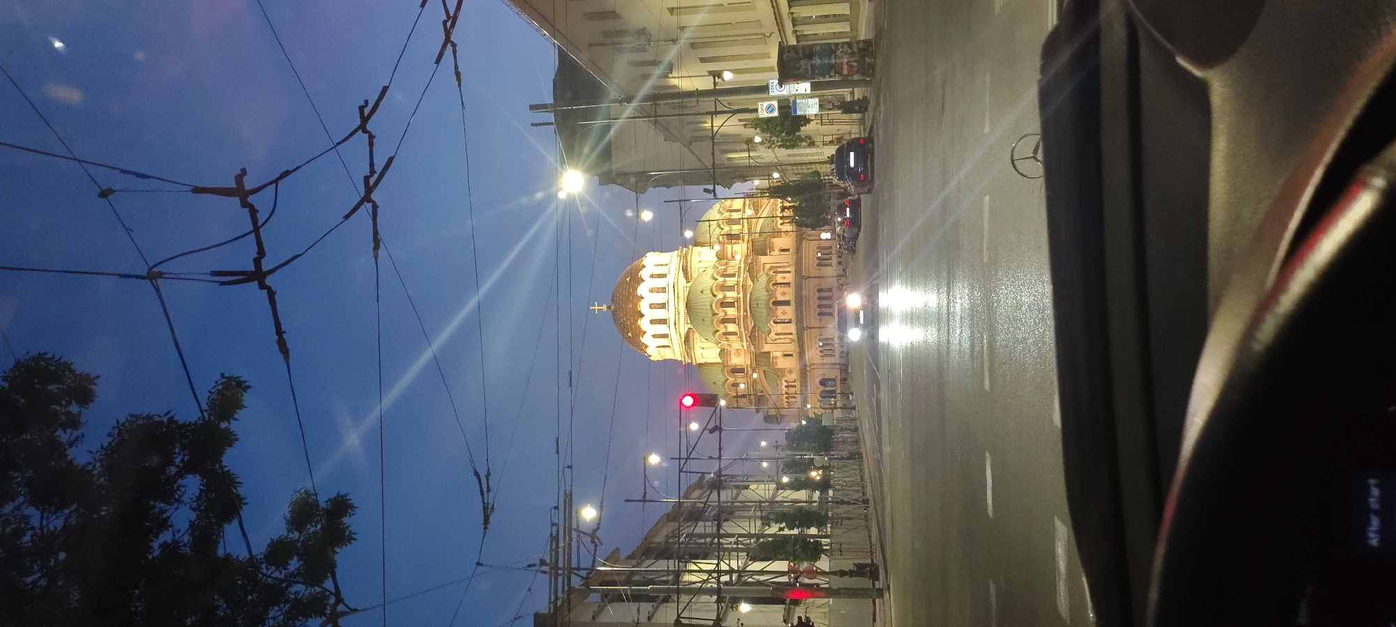 The image depicts a street scene in a city at dusk, with a large, illuminated cathedral in the background. The cathedral features a golden dome topped with a cross and several green domes, with its facade lit up in a warm yellow light. The sky above is a deep blue, with some clouds visible. Overhead wires and streetlights are prominent in the foreground and mid-ground, suggesting an urban environment. Several cars are on the wet street, some with their headlights on, indicating traffic. The view appears to be from inside a vehicle, as the dashboard and steering wheel are partially visible in the lower part of the frame. The overall impression is a view of an iconic landmark at the beginning of the evening.