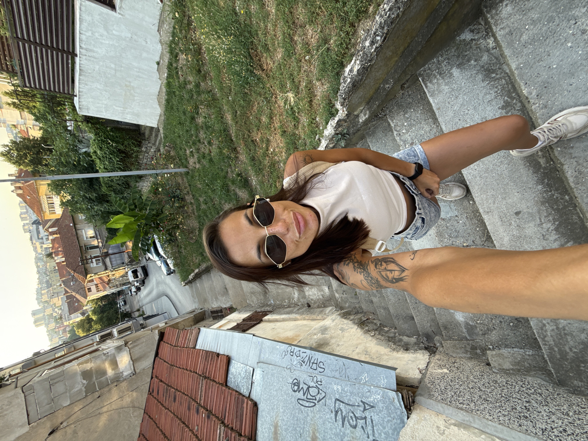 A woman with long brown hair and sunglasses is taking a selfie while descending a stone staircase in Pleven, Bulgaria. She is wearing a white tank top, denim shorts, and white sneakers. The staircase is made of concrete and appears to be quite old, with some graffiti on the adjacent wall. In the background, a street with parked cars and buildings can be seen. The overall scene suggests an urban environment.