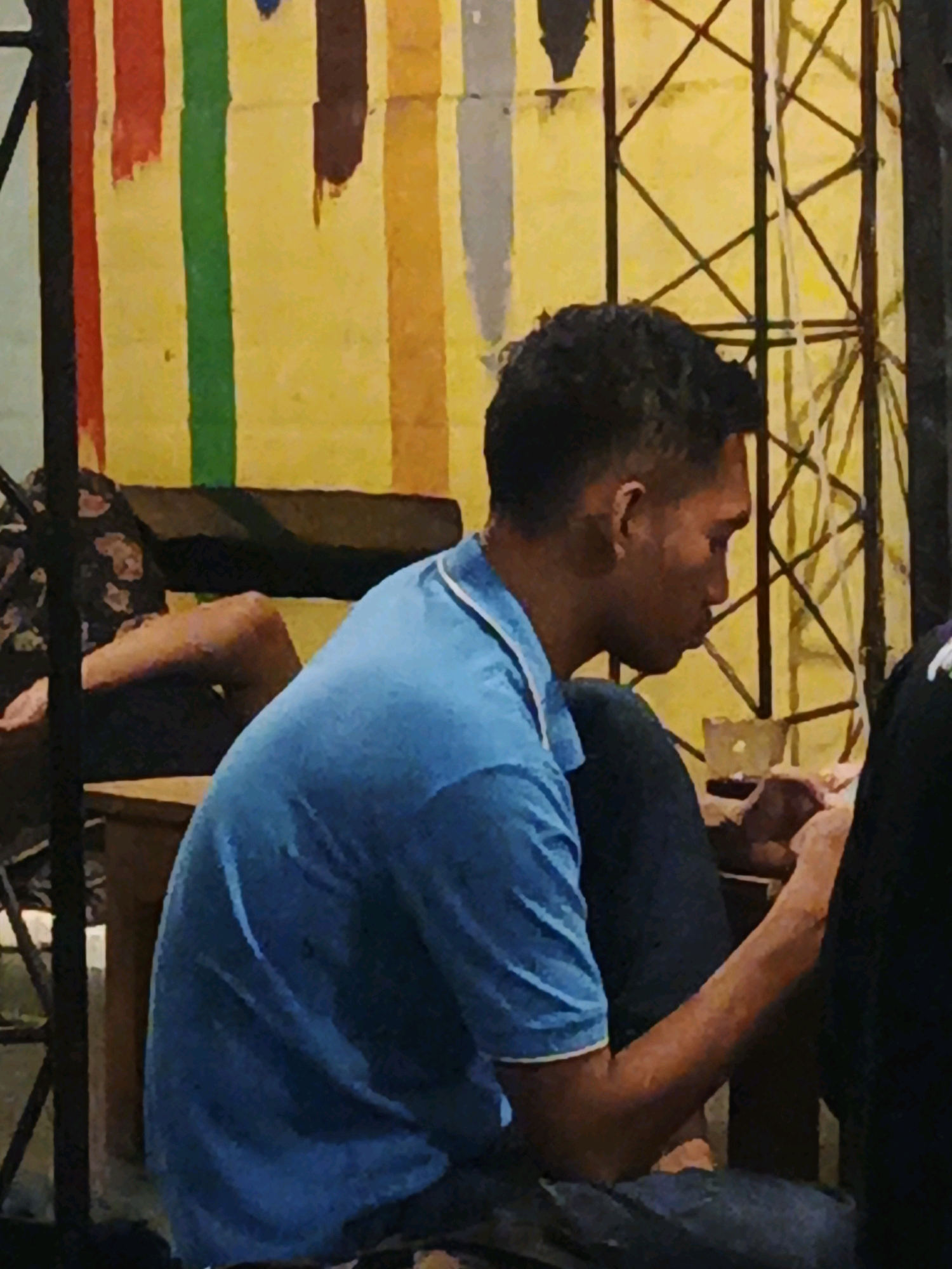 In the city of Ketegan, Indonesia, a medium shot captures a young man in a blue shirt with a white collar, seated and looking down. His dark hair is styled short, with a fade on the sides. He is positioned in the foreground, facing right, with his arms and hands visible. To his left and slightly behind him, another person is seated with their legs crossed, wearing a dark, patterned garment. The background features a wall painted with vertical stripes of various colors, including red, green, yellow, brown, and orange, set against a textured yellow backdrop. Overlapping the background is a dark, geometric metal structure. The scene appears to be indoors or under a covered area.