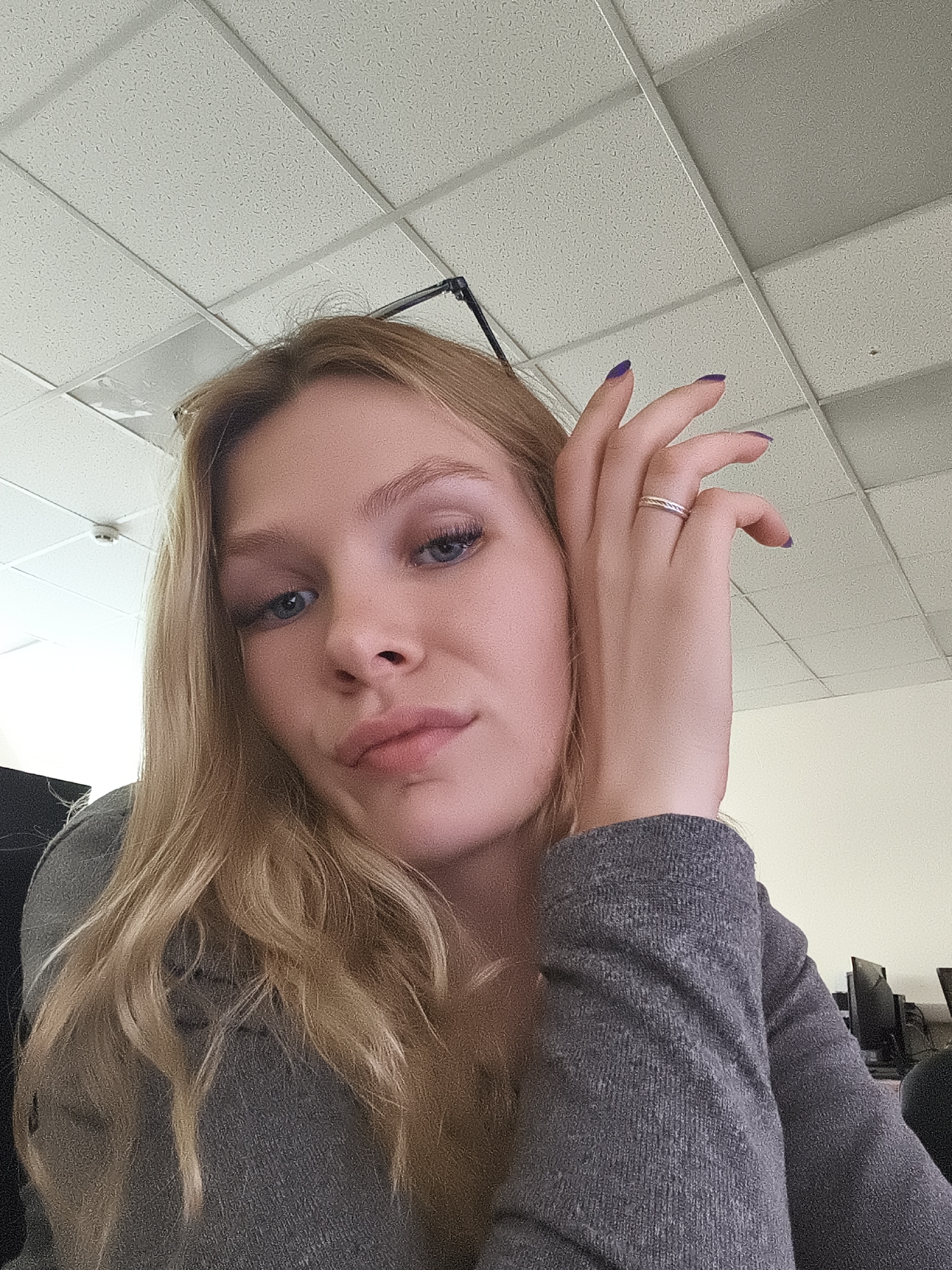A young woman with blond wavy hair and blue eyes poses for a selfie in what appears to be an office environment. She is wearing a gray long-sleeved top and a ring on her right ring finger. Her nails are painted purple. The background features a standard drop ceiling with square tiles, and glimpses of computer monitors suggest a workspace. The location within the city of Unknown, Unknown, is not discernible from the image.