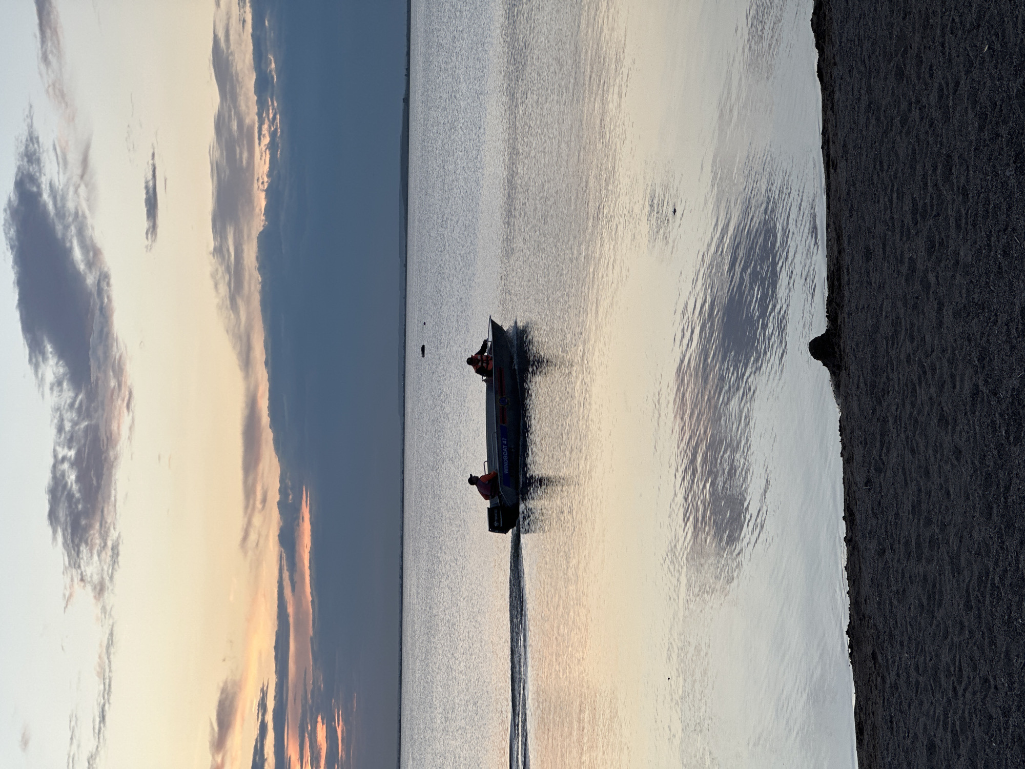 A small motorboat with two occupants is navigating a calm body of water at sunset. The boat, labeled "WINDBOAT 47," is moving from left to right, leaving a wake behind it. The sky is painted with soft hues of orange, pink, and blue, with scattered clouds reflecting on the water's surface. The foreground consists of a dark, sandy shore. The scene is likely the Bukhtarma Reservoir near Kokshetau, Kazakhstan, a popular recreational area.