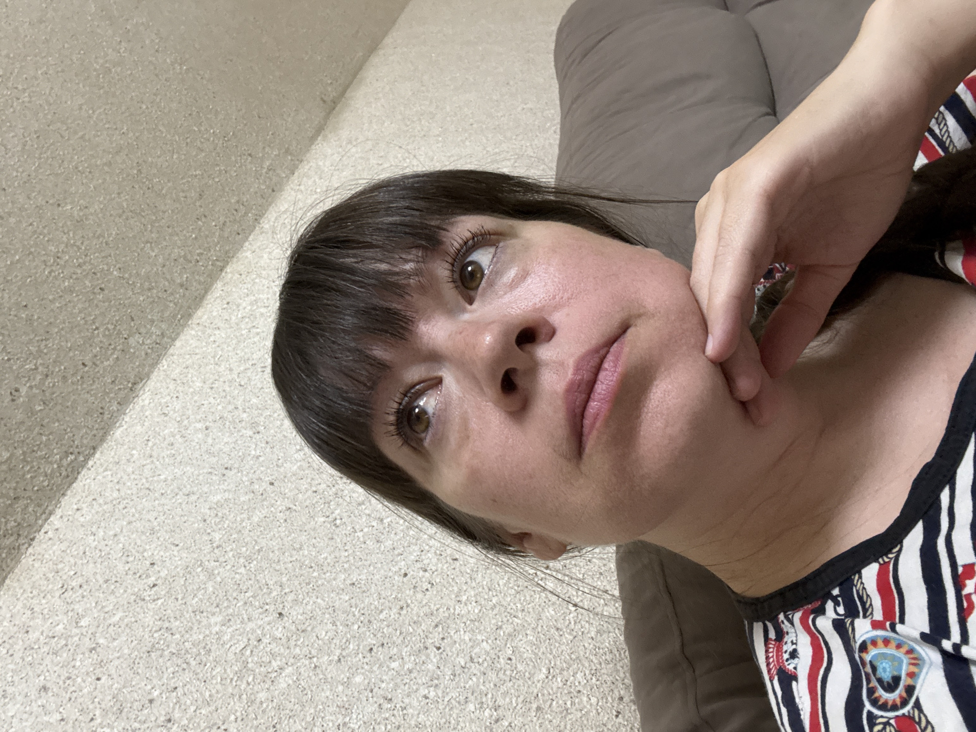 The image shows a close-up, high-angle shot of a woman with brown hair and bangs, looking upwards. Her right hand is resting on her chin. She is wearing a striped shirt with a black collar and a colorful heart-shaped emblem on the left side of her chest. The background appears to be a textured wall and a brown couch. The scene does not provide any information to determine its location in the city of Ветлужский, Russia.