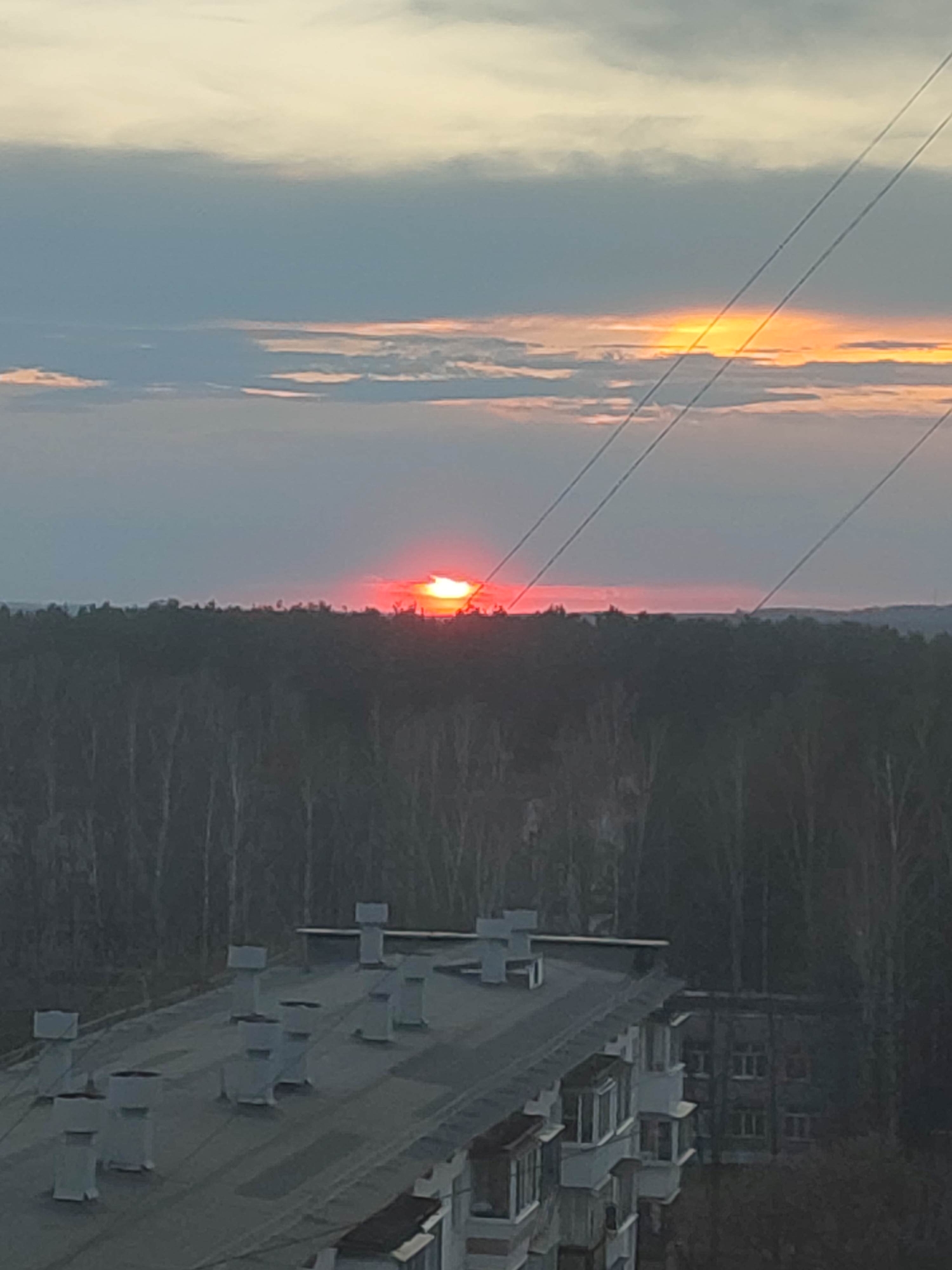 The image depicts a sunset over the city of Chaikovsky, Russia, with a building in the foreground and a forest in the mid-ground. The sky is a mix of orange, red, and grey clouds, with the sun visible on the horizon. Three electrical wires are running across the top right of the image. The building in the foreground has a grey roof with multiple white chimneys or vents. An adjacent dark building is also seen. The forest behind the building is dark green, indicating dense vegetation. The scene appears to be taken from an elevated position, such as a higher floor of a building.