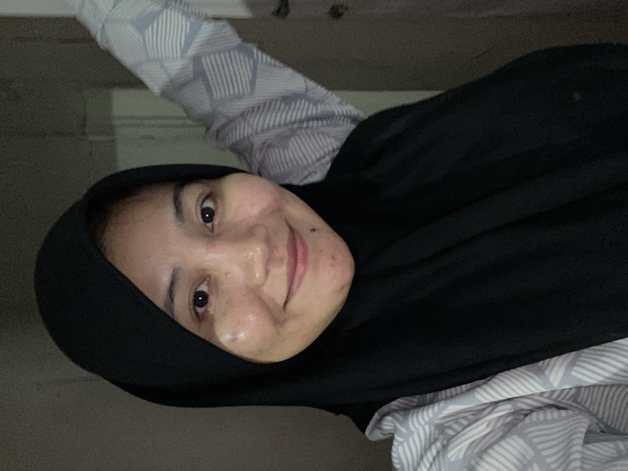 A medium shot captures a woman wearing a black hijab and a light-colored, patterned shirt, possibly in a home setting in Banjarbaru, Indonesia. Her head is tilted slightly, and she is smiling gently. The background is dimly lit with a textured wall and a glimpse of what appears to be a door or a closet. The lighting suggests the photo was taken indoors, likely in the evening or in a room with limited natural light. Her skin shows some texture, including what appears to be acne.