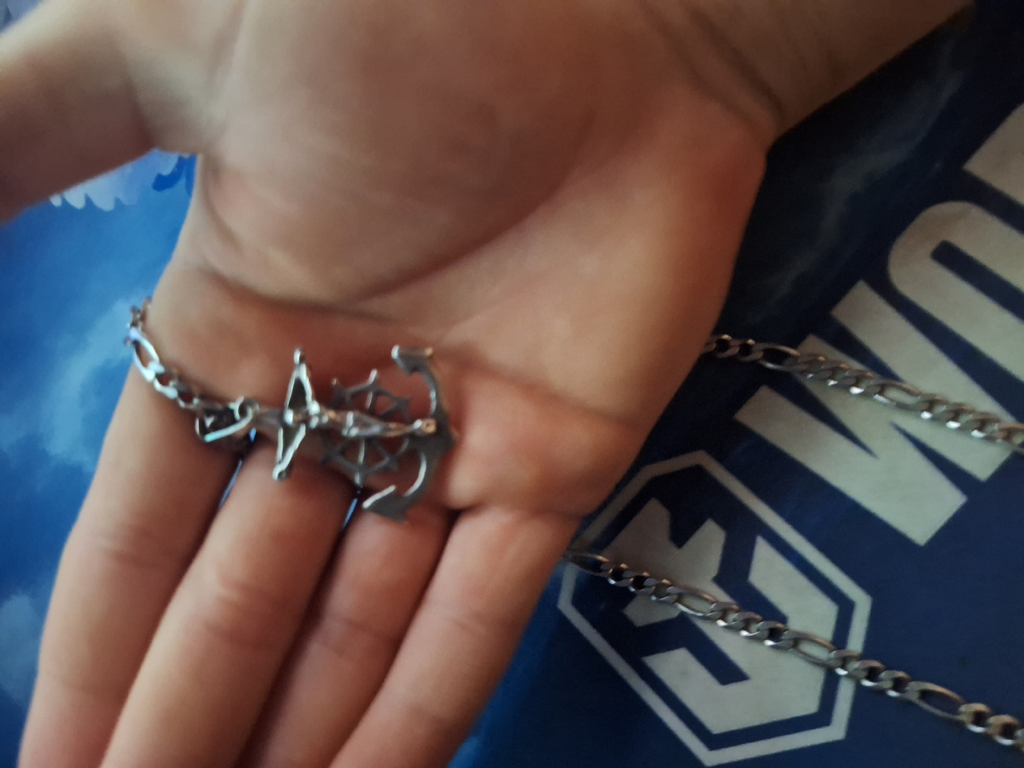 A close-up shot shows a person's hand holding a silver anchor pendant on a chain. The anchor is intricately designed, with a detailed wheel in the center. The hand is resting on a blue surface with a white graphic, possibly a logo or design. The background is out of focus, but appears to be a sky with some blue and white shapes. The lighting suggests it could be daytime, but the overall mood is neutral. The image's focus is on the jewelry. There is no visible text in the image.