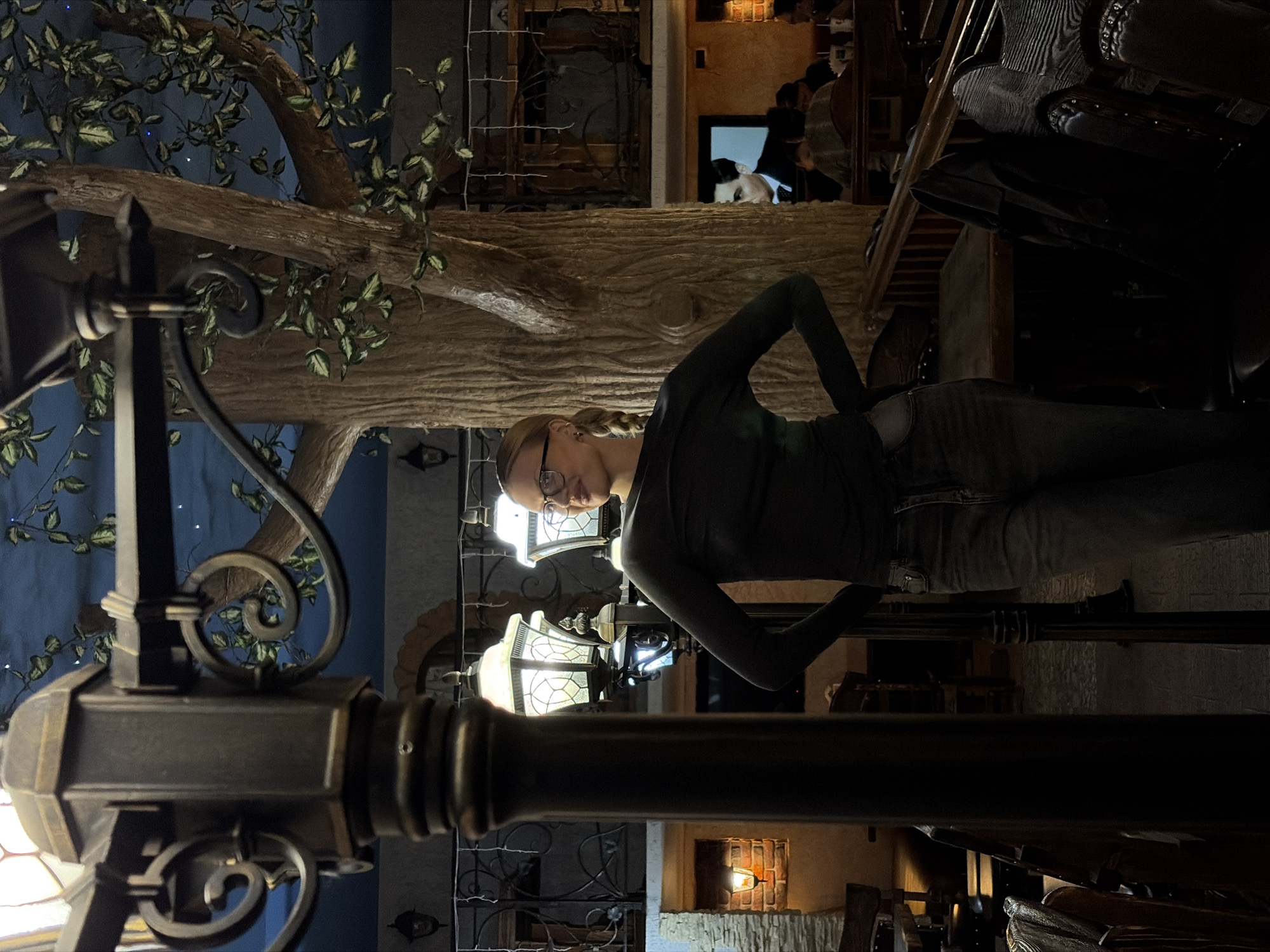 A woman wearing glasses, a dark long-sleeved shirt, and jeans stands sideways in an indoor setting, likely a restaurant in Chaikovsky, Russia. She poses with her hand on her hip. Behind her is an artificial tree with leaves, a wall with arched decorations, and other people in the background. A metal lamppost is in the foreground, partially obstructing the view. The orientation of the image is rotated to the left.