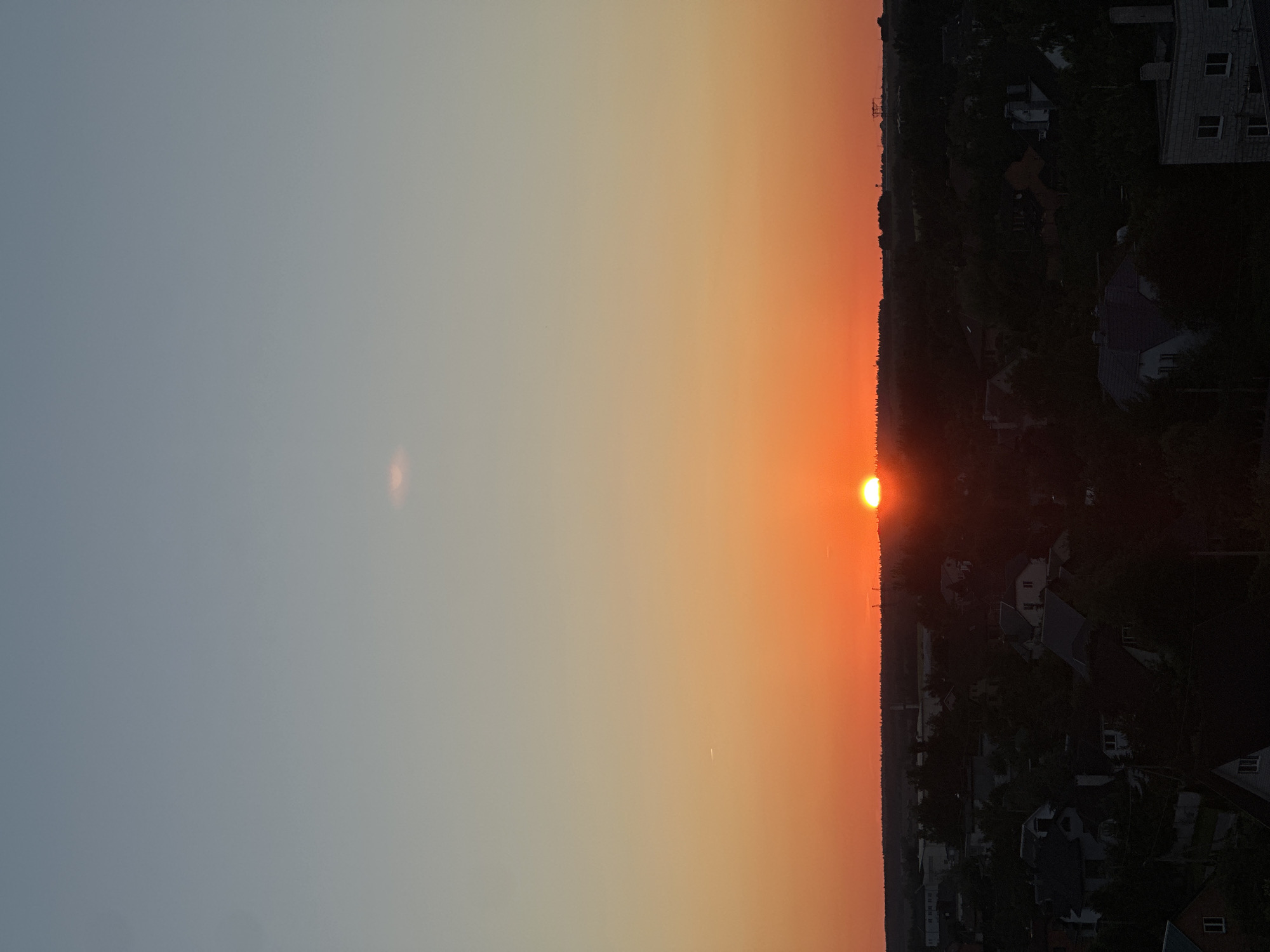 The image depicts a sunset over a suburban area in the vicinity of Шчомысліцкі сельскі Савет, Belarus. The sky transitions from a pale blue at the top to a vibrant orange hue near the horizon, with the sun appearing as a bright, round disc partially obscured by the silhouette of trees and buildings. A faint, wispy cloud is visible in the upper portion of the sky. The foreground shows a cluster of houses and dense foliage, indicating a residential area. The overall scene is one of a peaceful evening with the natural light of the setting sun casting long shadows over the landscape.