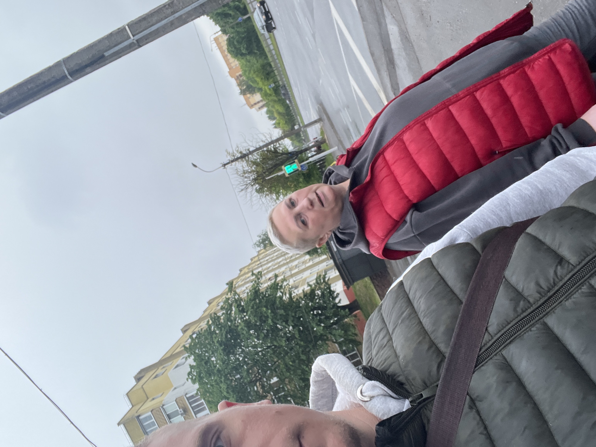 This image is a wide-angle selfie featuring two individuals outdoors on an overcast day, depicting an urban street scene in Minsk, Belarus.
The male subject, partially visible on the left side of the frame, is taking the photograph. His left eye, part of his nose, mouth, and chin are visible. He has short, light-colored hair and light-colored eyes. He is wearing a light grey hooded sweatshirt layered under a dark olive green quilted puffer vest. A dark strap, possibly from a bag, crosses his chest. His gaze is directed towards the camera.
To the right of the male subject, a female individual is fully visible. She has light skin and short, light grey/blonde hair. She is wearing a dark grey hooded sweatshirt beneath a bright red quilted puffer vest. Her expression is neutral as she also looks towards the camera.
The foreground shows wet pavement, indicating recent precipitation. Immediately behind the subjects, a multi-lane asphalt road is visible, featuring a white-striped pedestrian crosswalk. A green traffic light is illuminated on a pole positioned to the right of the female subject, adjacent to the road. Further right, a tall concrete utility pole with metal bands is present. In the midground and background, multi-story residential buildings, primarily with yellow and white facades, are visible. Lush green trees are scattered throughout the scene, bordering the buildings and the street. A dark-colored vehicle is discernible on the road in the distance. The sky is uniformly grey and cloudy.
