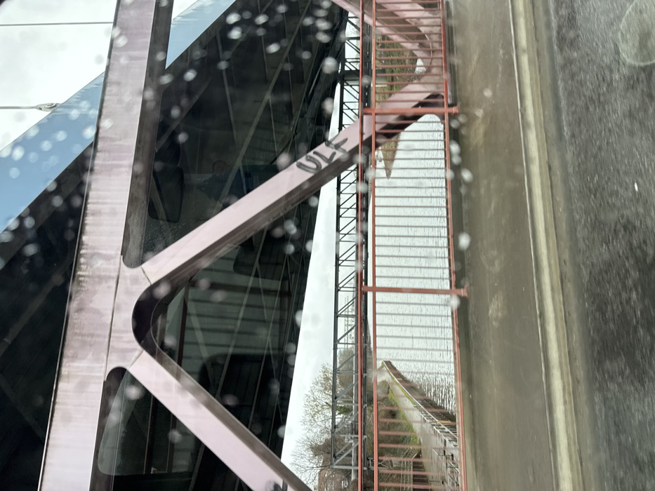 The image depicts a view from inside a vehicle looking out onto the Rhein-Herne Canal in Bottrop, Germany, specifically featuring the Rheinbabenbrücke (Rheinbaben Bridge) carrying the B224. In the immediate foreground, a blurred dark surface, likely the vehicle's dashboard or part of the road, is visible. The camera lens or window is covered in numerous raindrops, creating a speckled, out-of-focus effect across the entire image.
The prominent subject is the large, weathered reddish-brown metal truss structure of the Rheinbabenbrücke, which arches over the canal. Its robust beams and supports are visible from below the bridge deck. Black graffiti, spelling "ULF", is visible on one of the vertical beams on the right side of the main bridge structure. Below the main bridge deck, a red-orange painted metal railing with vertical bars runs along the edge of the bridge, overlooking the canal.
Below the Rheinbabenbrücke, the grey, rippled surface of the Rhein-Herne Canal flows. The left bank of the canal features a paved path running alongside a concrete or stone retaining wall, partially obscured by more red-orange vertical railings. On the far bank, sparse, leafless trees are visible against an overcast, light grey sky. In the mid-ground, spanning the canal parallel to the Rheinbabenbrücke, is another, darker-colored metal truss bridge, likely a railway or industrial bridge, with a more intricate lattice work design. No people or active interactions are visible in the scene. The overall environment suggests an overcast, damp, possibly rainy day.