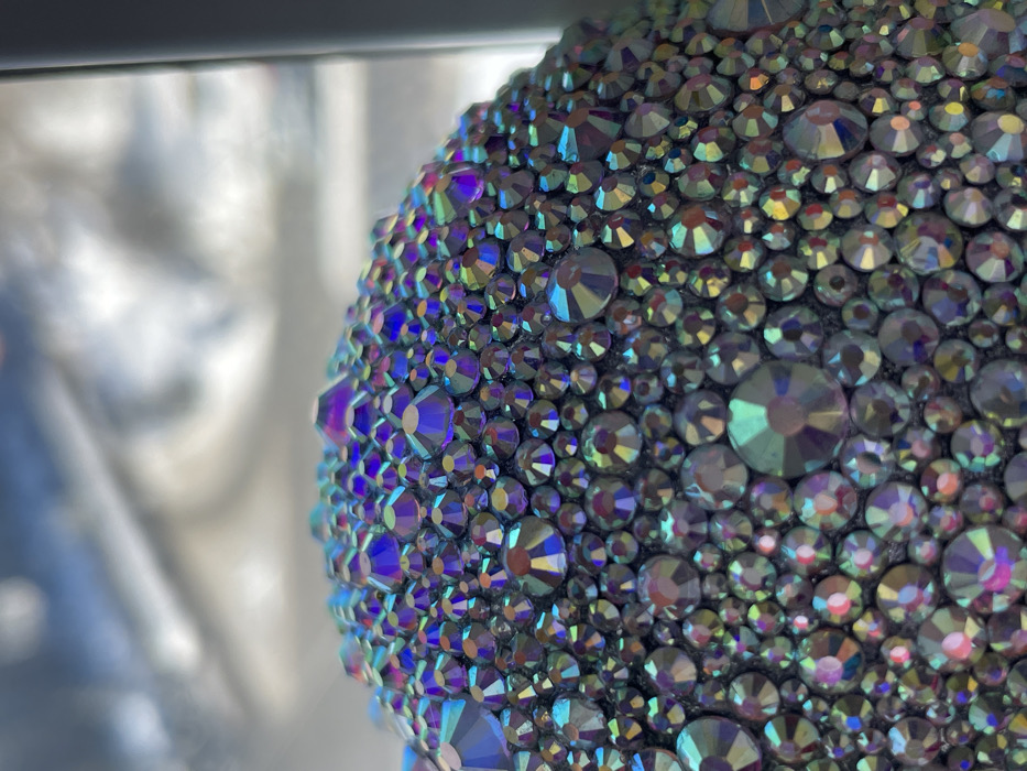 The image presents a close-up, high-detail view of a surface entirely covered with numerous iridescent rhinestones or crystals. These embellishments are densely packed together, varying significantly in size from very small to moderately large, and are affixed to a dark, likely black, underlying material.
Each rhinestone is faceted, catching and reflecting light in a vibrant, kaleidoscopic manner. The dominant colors reflected are rich blues, deep purples, emerald greens, and golden yellows, characteristic of an Aurora Borealis (AB) finish, which creates an ethereal, shimmering effect. The reflective qualities of the rhinestones give the surface a highly textured and sparkling appearance.
The object adorned with these rhinestones occupies the right and lower sections of the frame. The left side of the image is heavily blurred and out of focus, showcasing a soft bokeh background with indistinct shapes and varying levels of brightness. This blurred background suggests an indoor setting, possibly near a window or a light source, which contributes to the way the rhinestones brilliantly refract light. No specific activity is taking place; the image is a static, detailed shot of the embellished object. There is no visible text in the image.