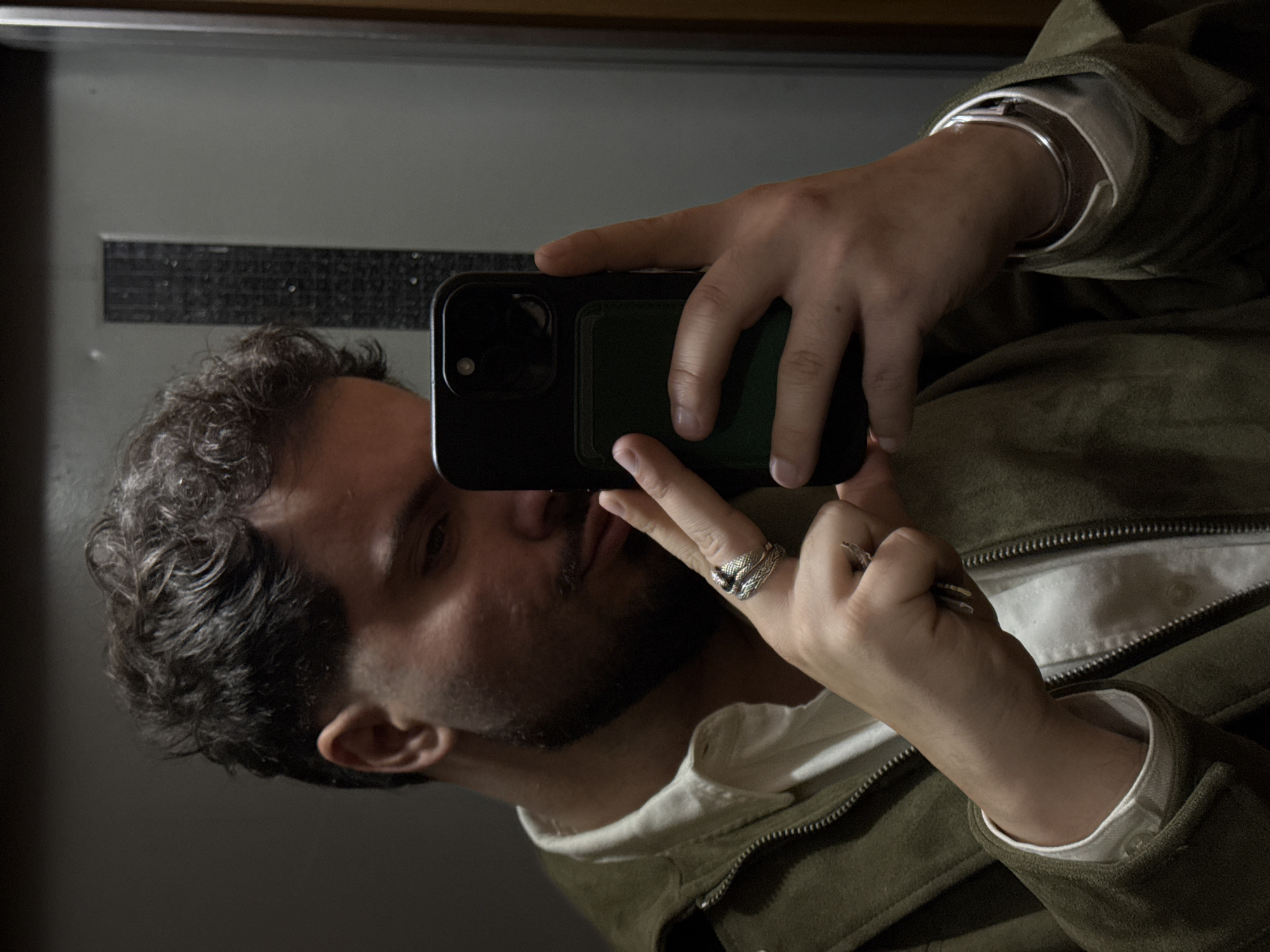 A male individual with dark, curly hair and a short, dark beard is depicted in a mirror selfie taken indoors in Vila Nova de Gaia, Portugal. He wears a white collared shirt underneath an unzipped olive-green jacket. His left hand holds a black smartphone, equipped with a dark green cardholder on its back, positioned to capture his reflection. The left hand displays a prominent silver-colored ring with a serpent design on the middle finger and another smaller ring on the ring finger. A silver-colored bracelet is visible on his left wrist beneath the jacket cuff. The background, visible in the reflection, consists of a grey-toned wall with a vertical, textured panel and a dark brown vertical element on the right, likely a door frame or architectural feature.