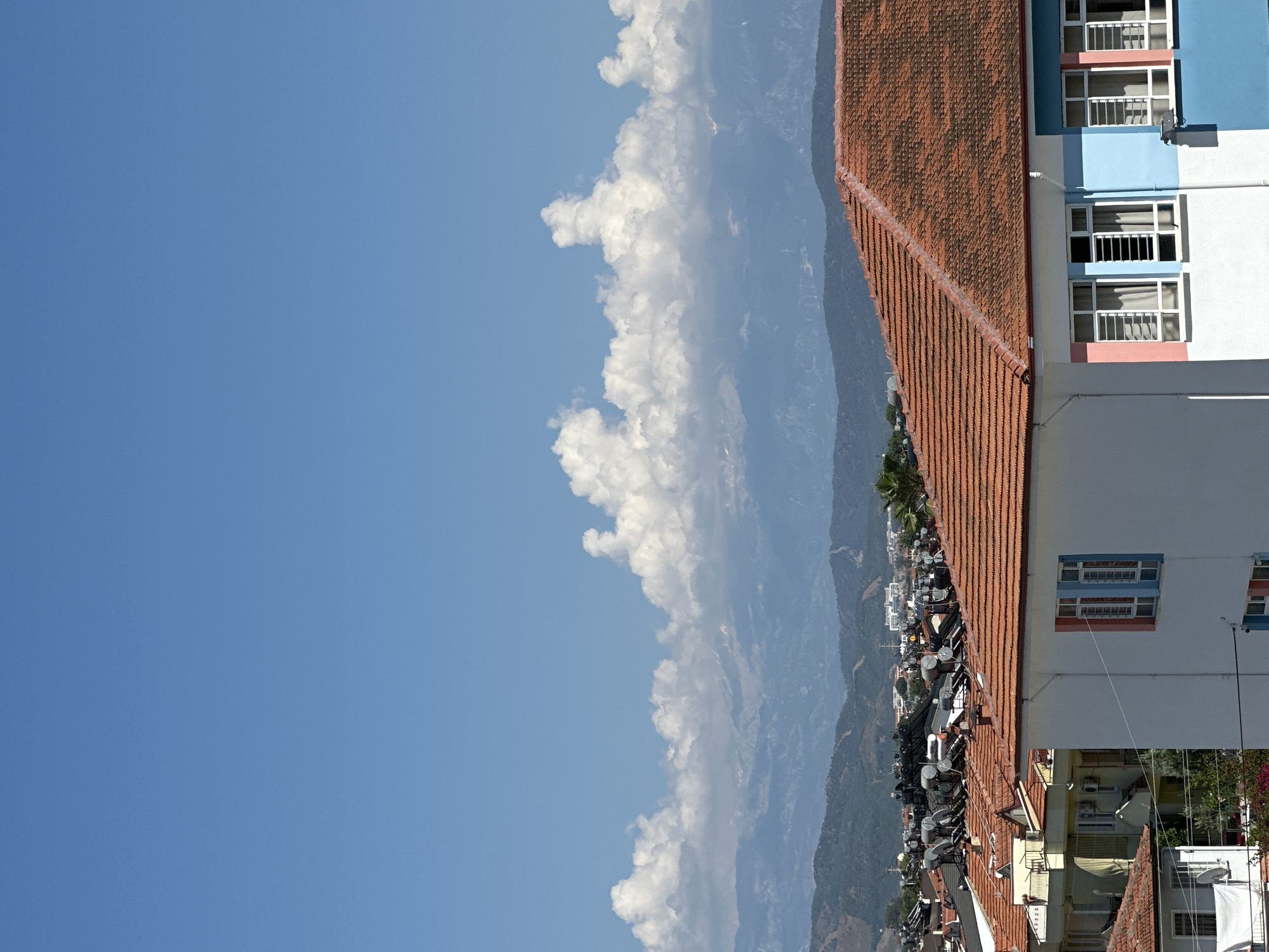A vertically-oriented photograph captures an urban scene in Fethiye, Turkey. The left side of the image showcases a blue sky with fluffy white clouds partially obscuring a view of distant mountains. Adjacent to the sky, a densely populated hillside descends, dotted with buildings. The right side of the photograph focuses on the roofs of several buildings, covered in orange clay tiles. The side of a building is also visible, featuring a facade with a light blue and white exterior and windows with white frames. The location is likely within Fethiye, Turkey, based on the visible architectural elements and the hilly terrain.