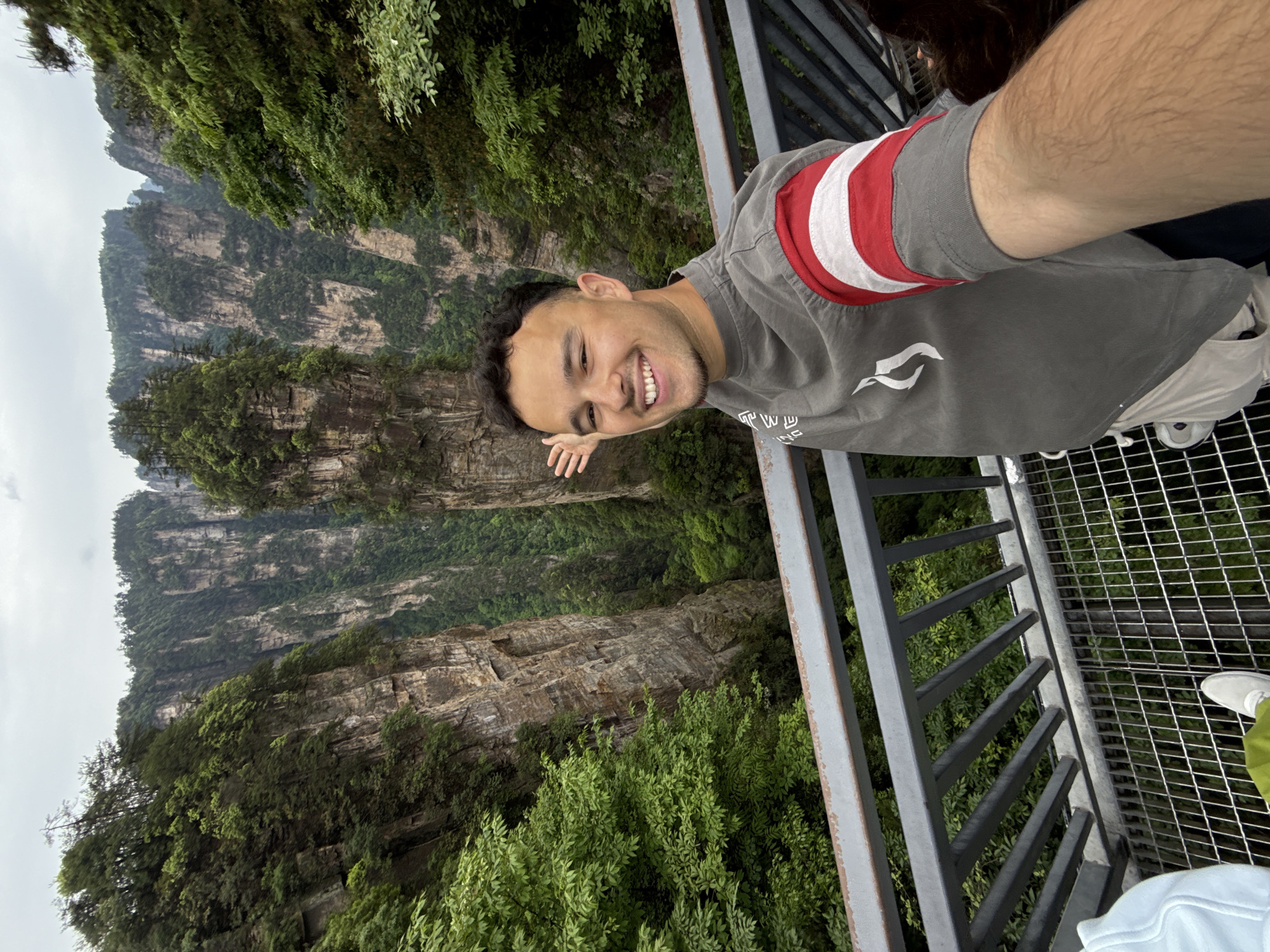 A medium-shot photo shows a smiling young man standing on an observation deck with metal railings, likely a selfie. He is wearing a gray t-shirt with a red and white stripe design on the sleeve, and light-colored shorts or pants. He is positioned on the right side of the frame with his face towards the upper right corner. His right arm is extended towards the middle left of the frame.
Behind him, the landscape consists of tall, vertical rock formations covered in lush green vegetation, suggesting a mountainous or canyon area. The sky is overcast, creating a soft, diffused light.
The location is suggested to be in the city of Luoguta, China, based on the prompt's instructions.